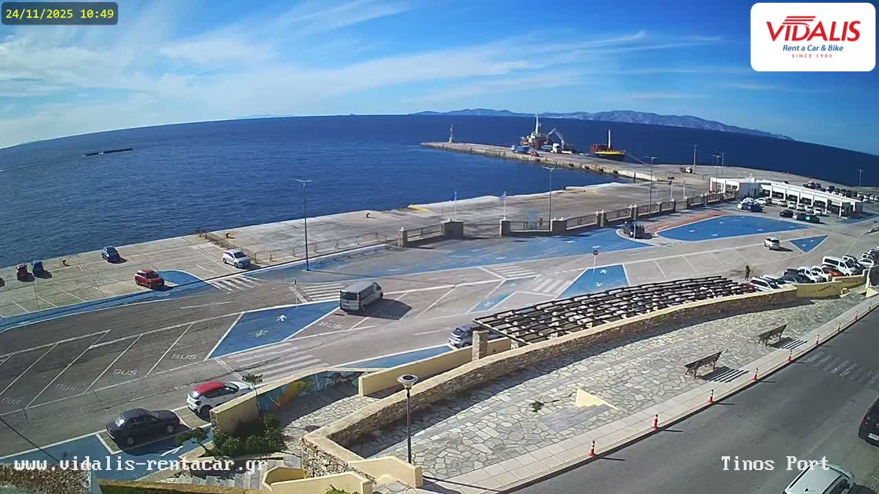 A vibrant port is seen on a clear, sunny day, featuring a calm blue sea adjacent to a concrete pier with parked vehicles, marked lanes, a lighthouse, and a large vessel, all framed by distant mountains under a blue sky with scattered clouds.