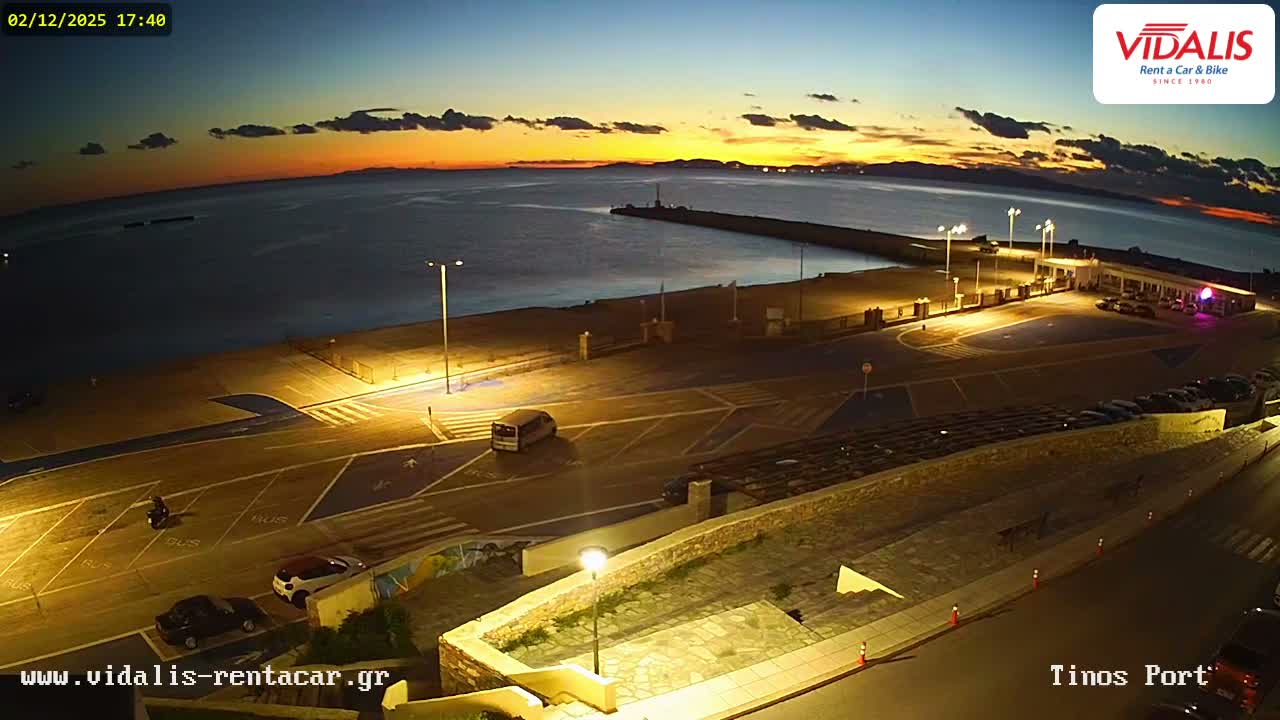 A wide-angle view captures a clear twilight scene at a bustling port, featuring a long pier extending into the sea, an illuminated terminal with parked cars, and a few vehicles moving on the roads under a vibrant sunset sky.