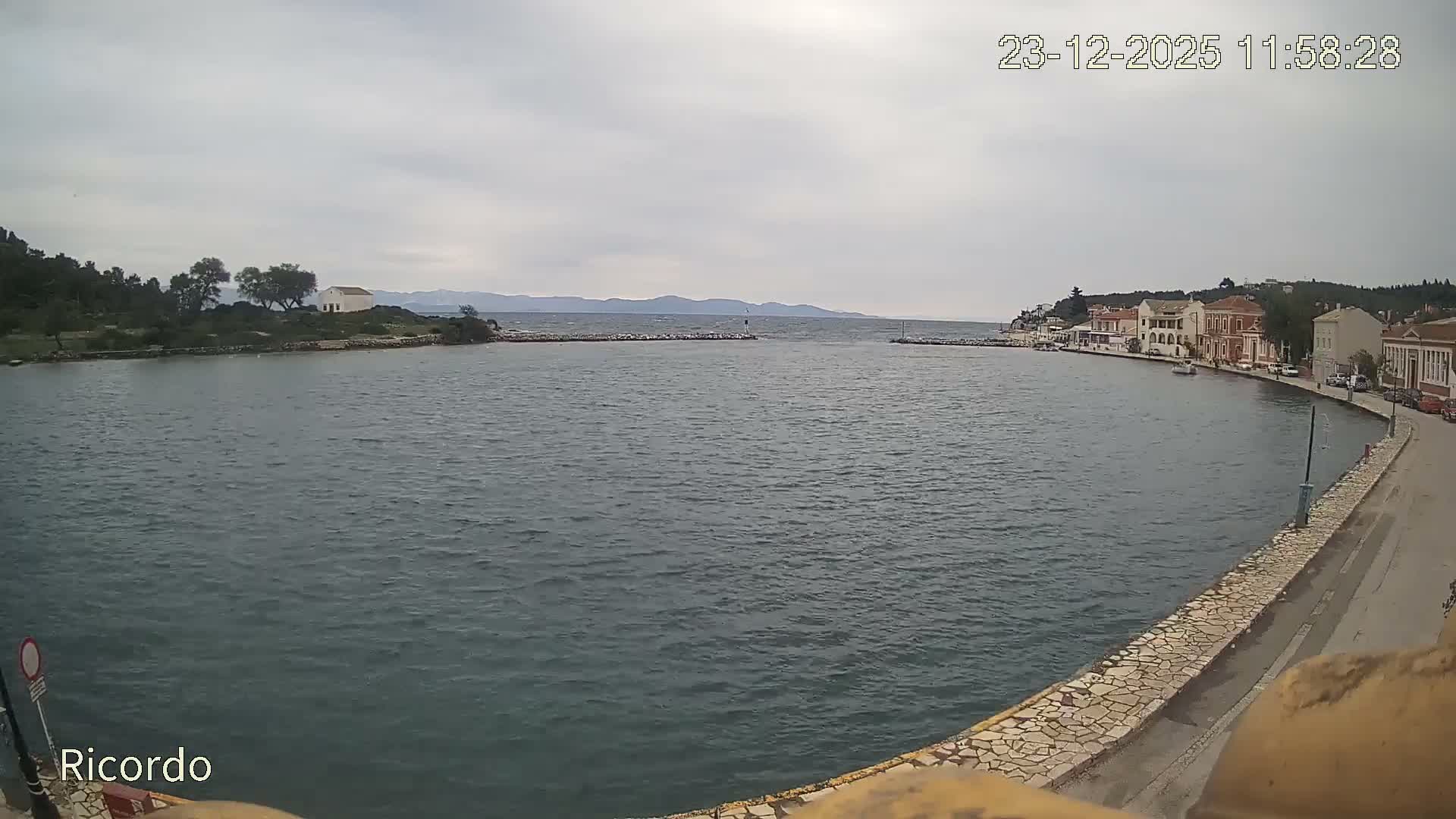 A coastal town with buildings lining a curving waterfront promenade overlooks a tranquil bay under an overcast sky, with distant hills visible beyond the sea entrance.