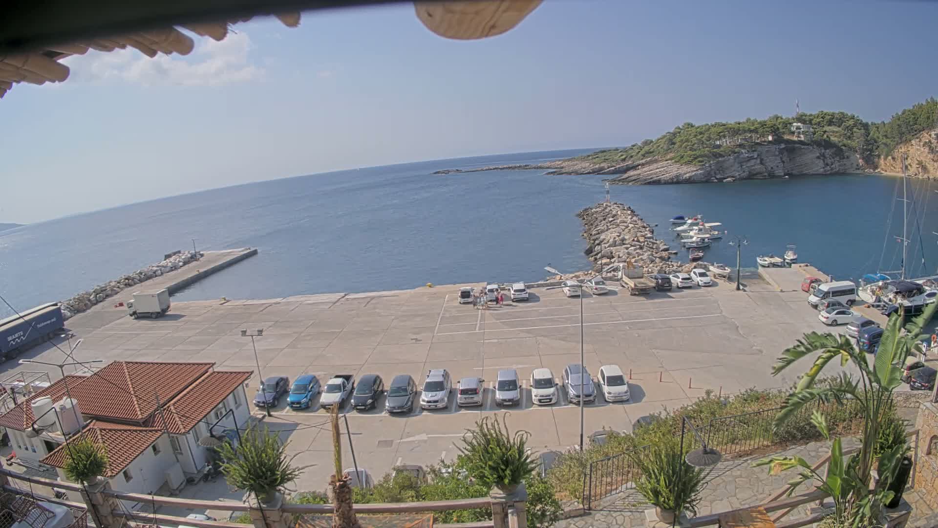 A sunny day at a harbor shows a row of cars parked in a lot overlooking a calm sea with several boats moored.