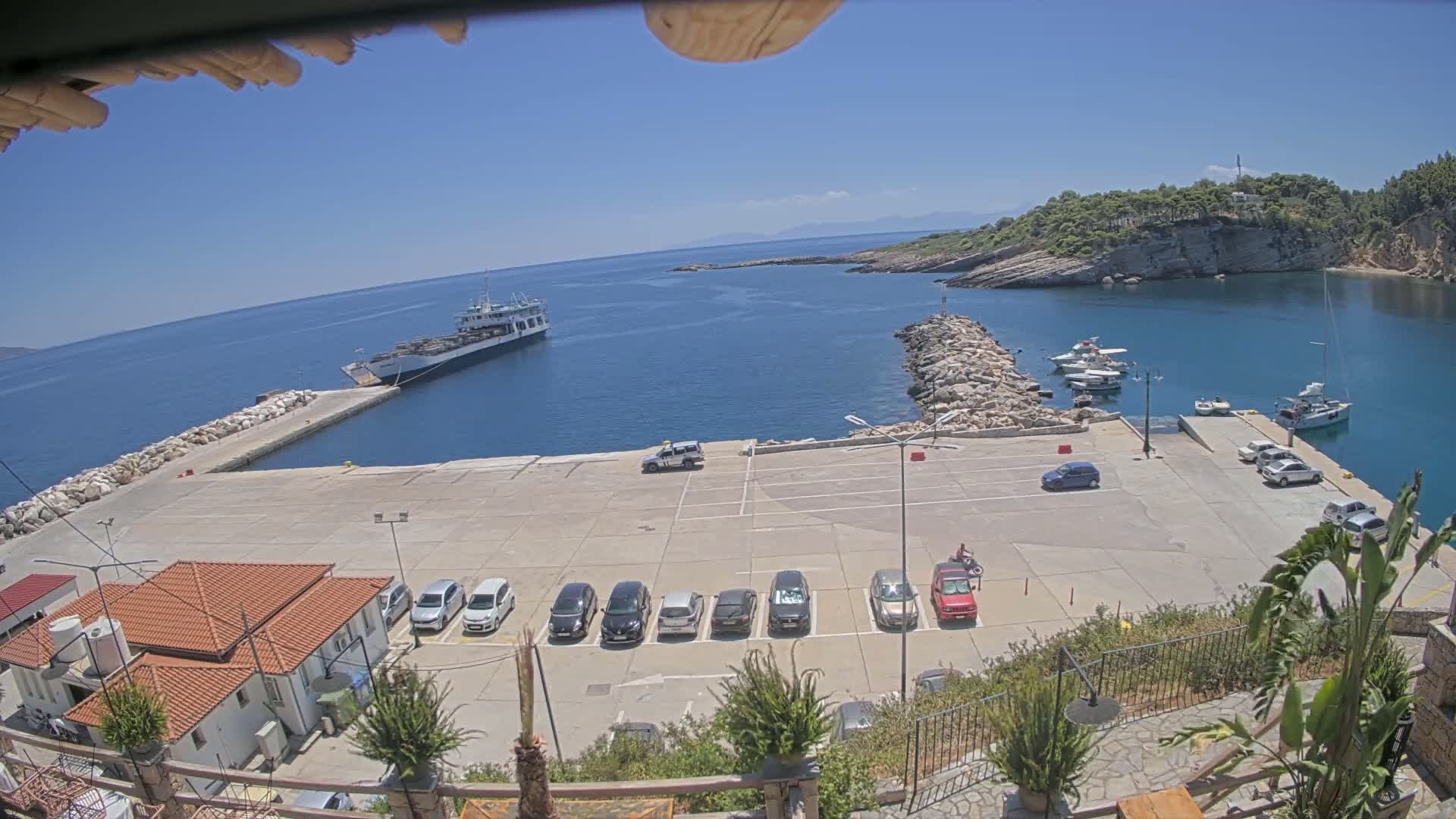 A sunny day at a harbor shows a row of cars parked in a lot overlooking a calm sea with several boats moored.