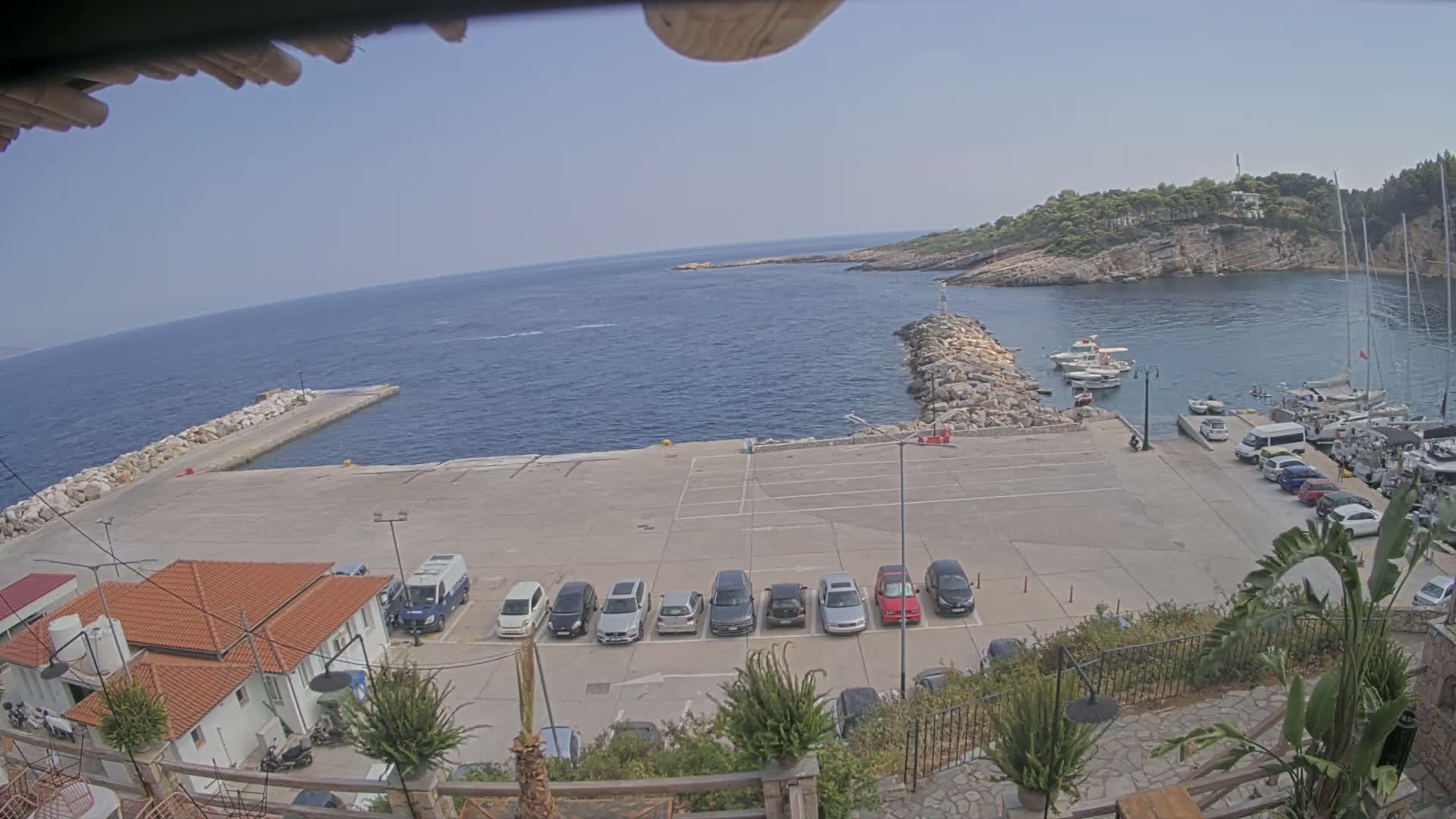 A sunny day at a harbor shows a row of cars parked in a lot overlooking a calm sea with several boats moored.