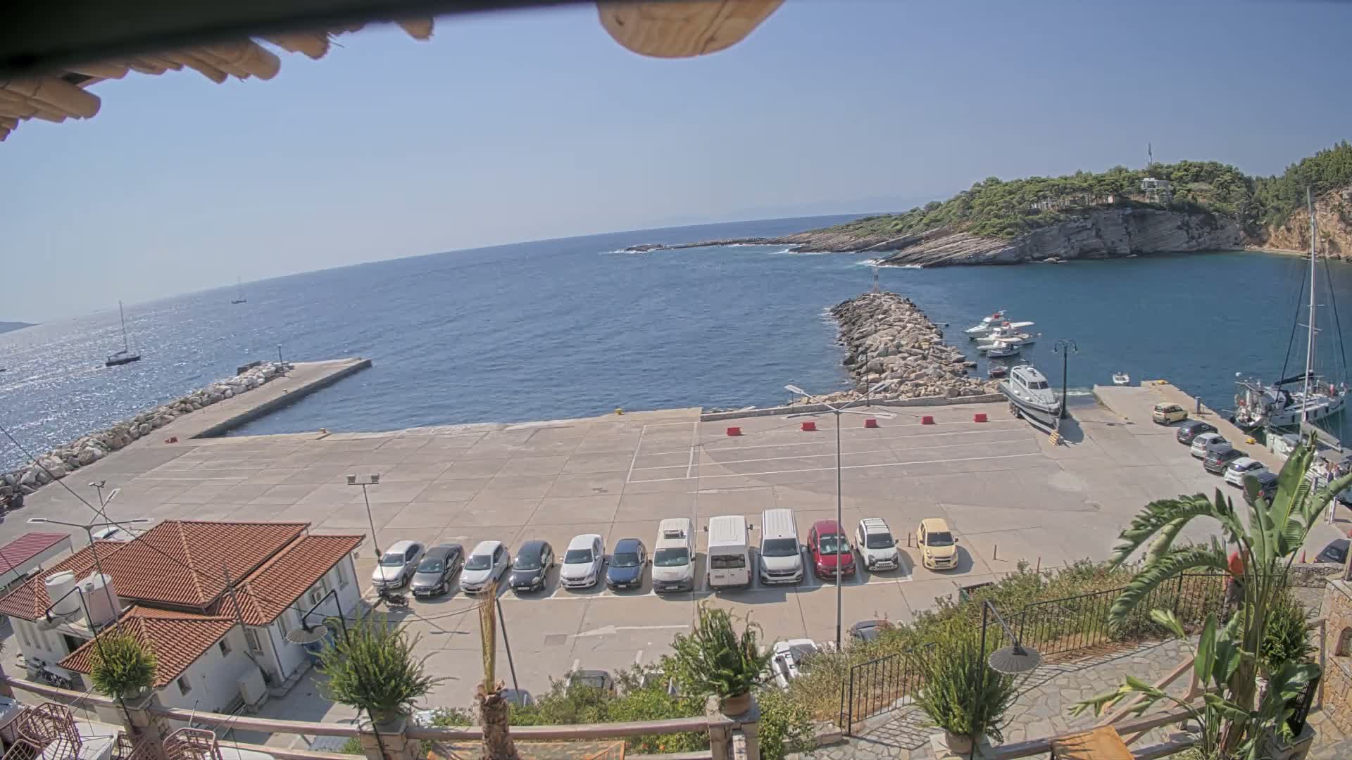 A small harbor with a row of cars parked on a concrete area, several boats moored in the water, and a rocky shore under a clear, sunny sky.