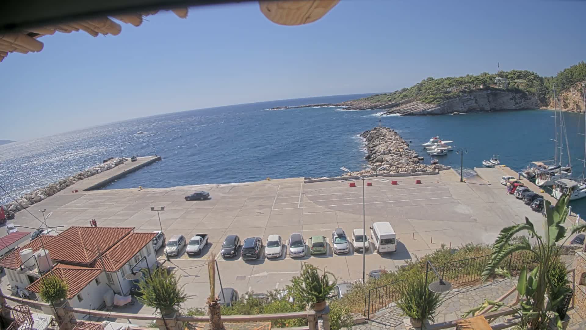 A sunny day shows a small harbor with a concrete parking lot full of cars, a stone jetty, several boats in the water, and a small building with a red tile roof.