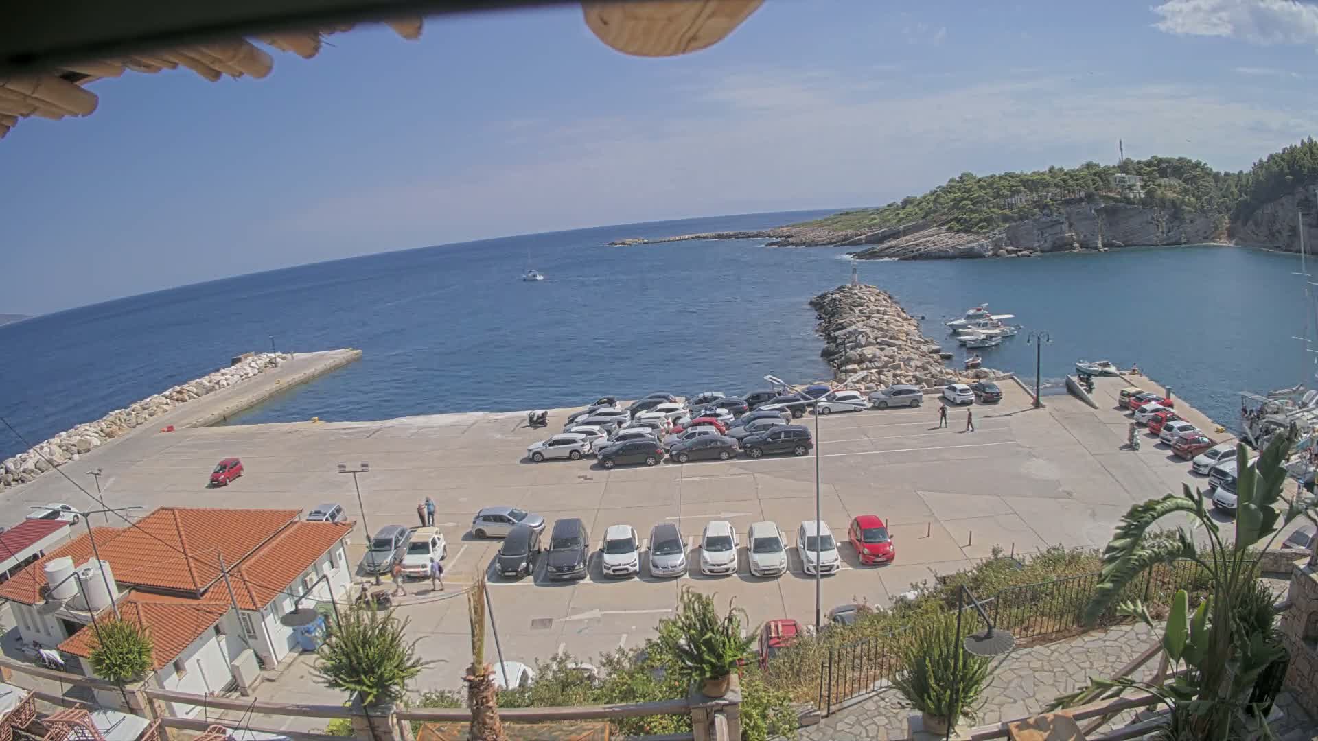 A sunny day shows a small harbor with a concrete parking lot full of cars, a stone jetty, several boats in the water, and a small building with a red tile roof.