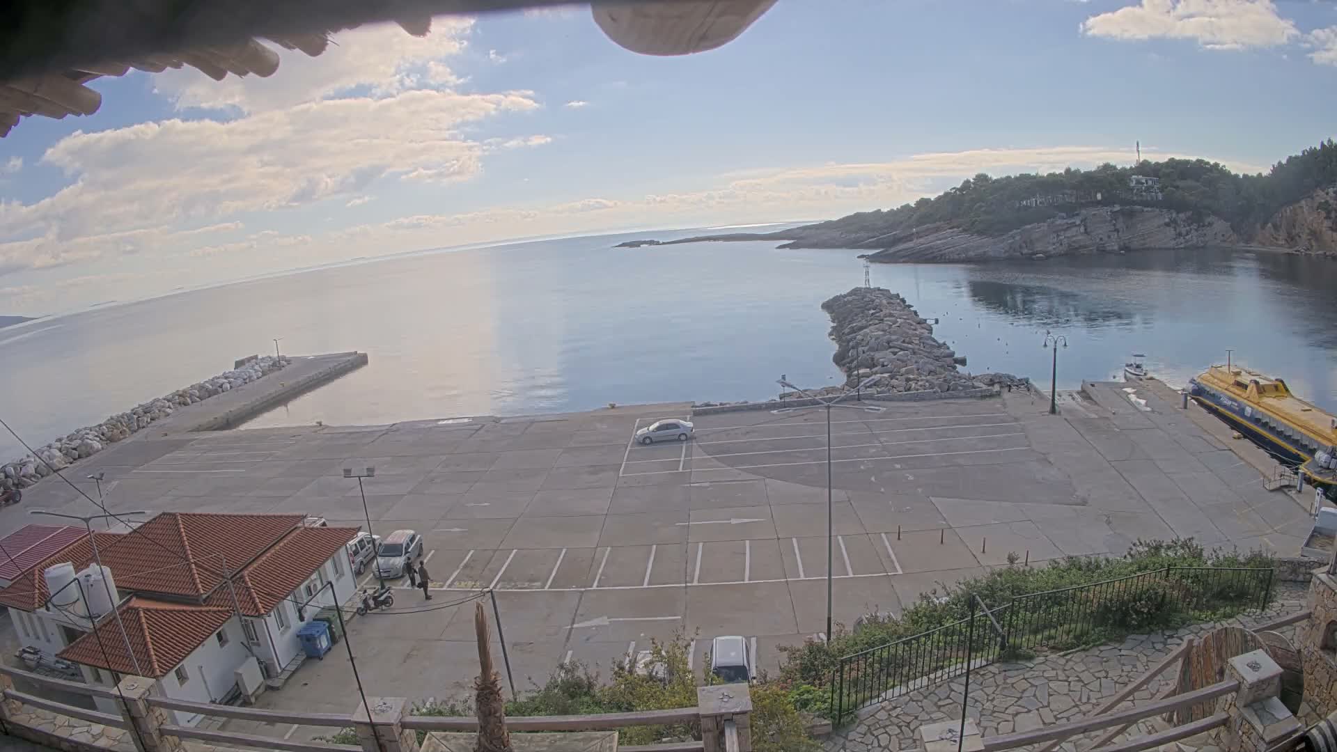 On a sunny, partly cloudy day, an elevated view captures a calm bay with two rock piers, a concrete parking lot and dock area featuring a large yellow and blue boat, and green hills rising in the distance.