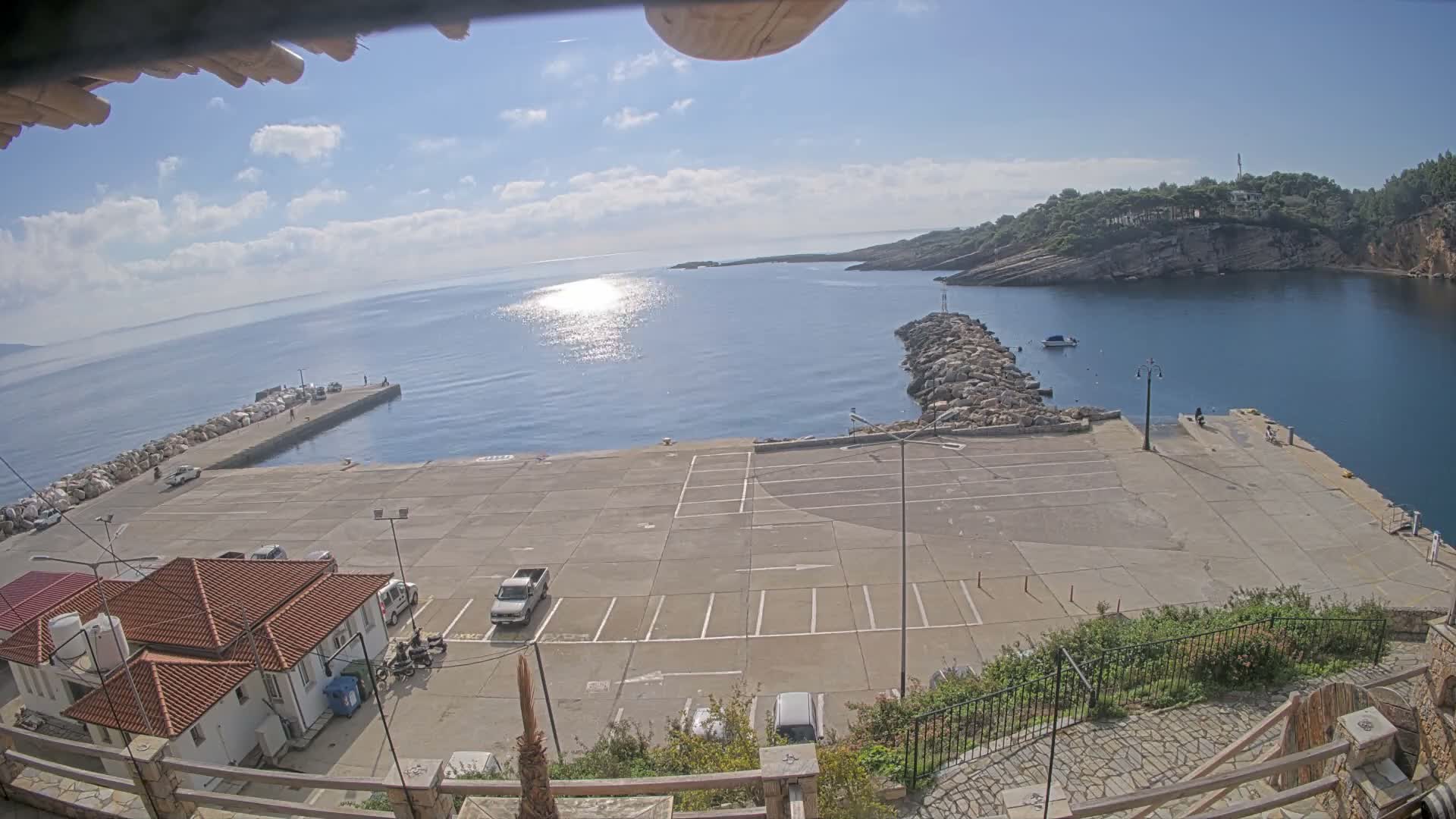 On a bright, partly cloudy day, a coastal bay with sun-dappled water is viewed from above, revealing a concrete pier, a rock breakwater, a few parked vehicles, and distant lush, rocky hills.