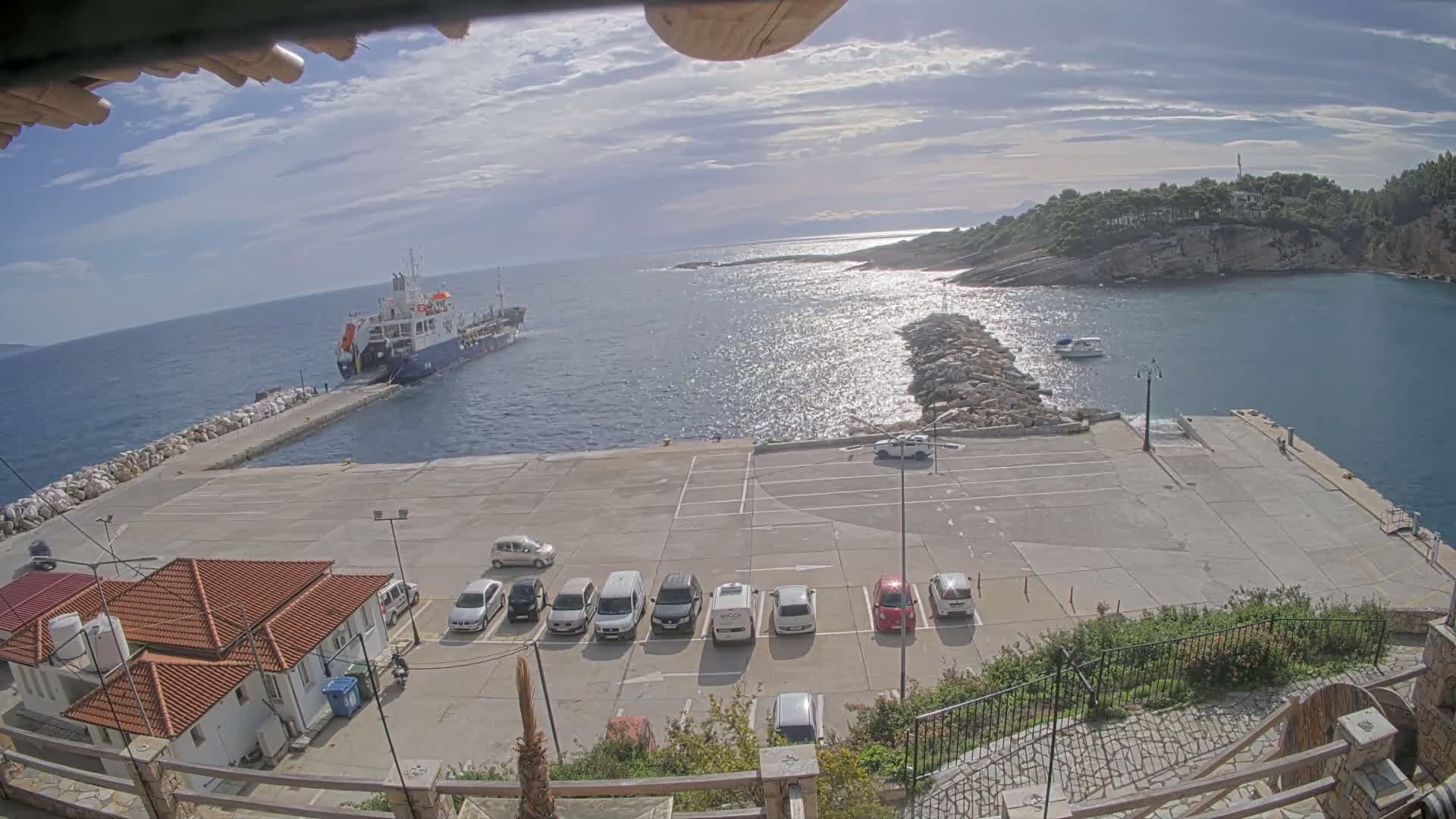 On a bright, partly cloudy day, a coastal bay with sun-dappled water is viewed from above, revealing a concrete pier, a rock breakwater, a few parked vehicles, and distant lush, rocky hills.