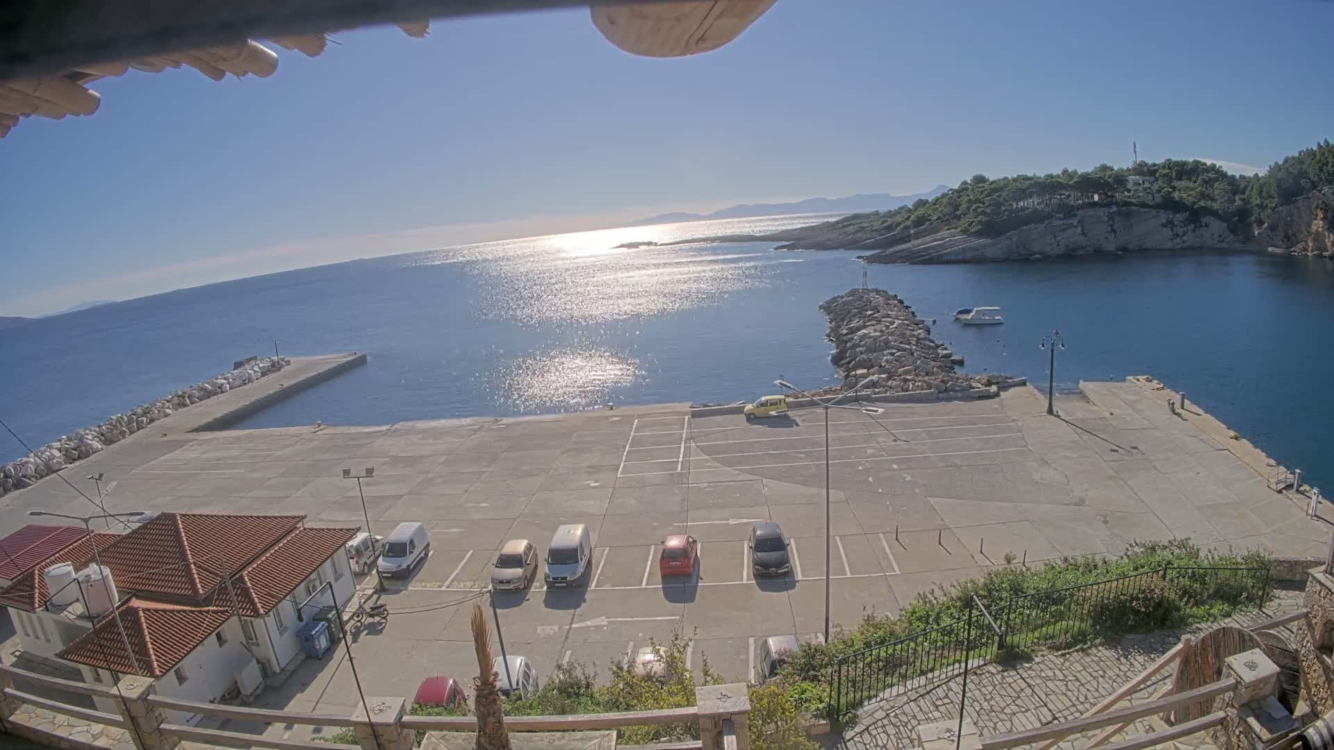 A bright sunny day illuminates a coastal scene featuring a concrete parking lot with several cars, traditional buildings, a long pier extending into calm blue waters where sunlight glints, and forested hills forming the distant shoreline under a clear sky.