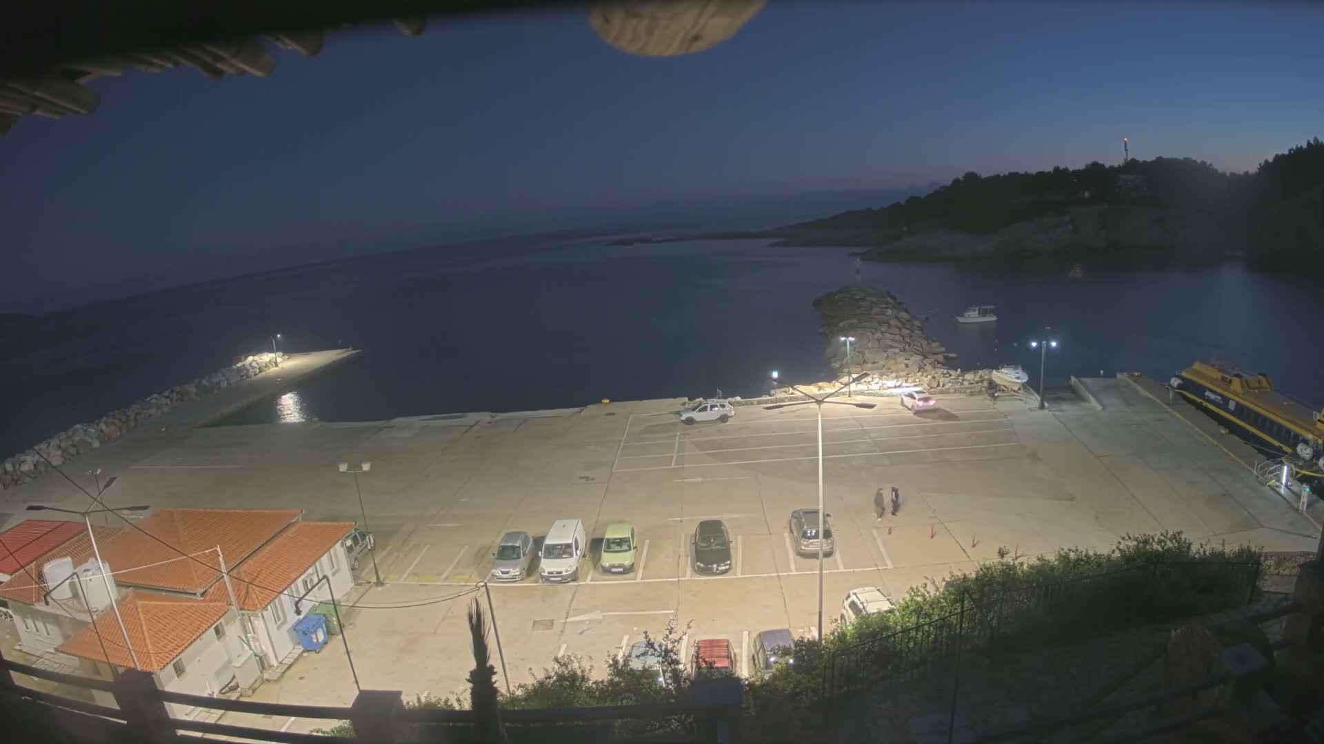 A clear night view from an elevated perspective shows a well-lit port area with a concrete dock, multiple parked cars, a few pedestrians, various boats including a large yellow ferry, and shoreline buildings, all under a dark, calm sky.