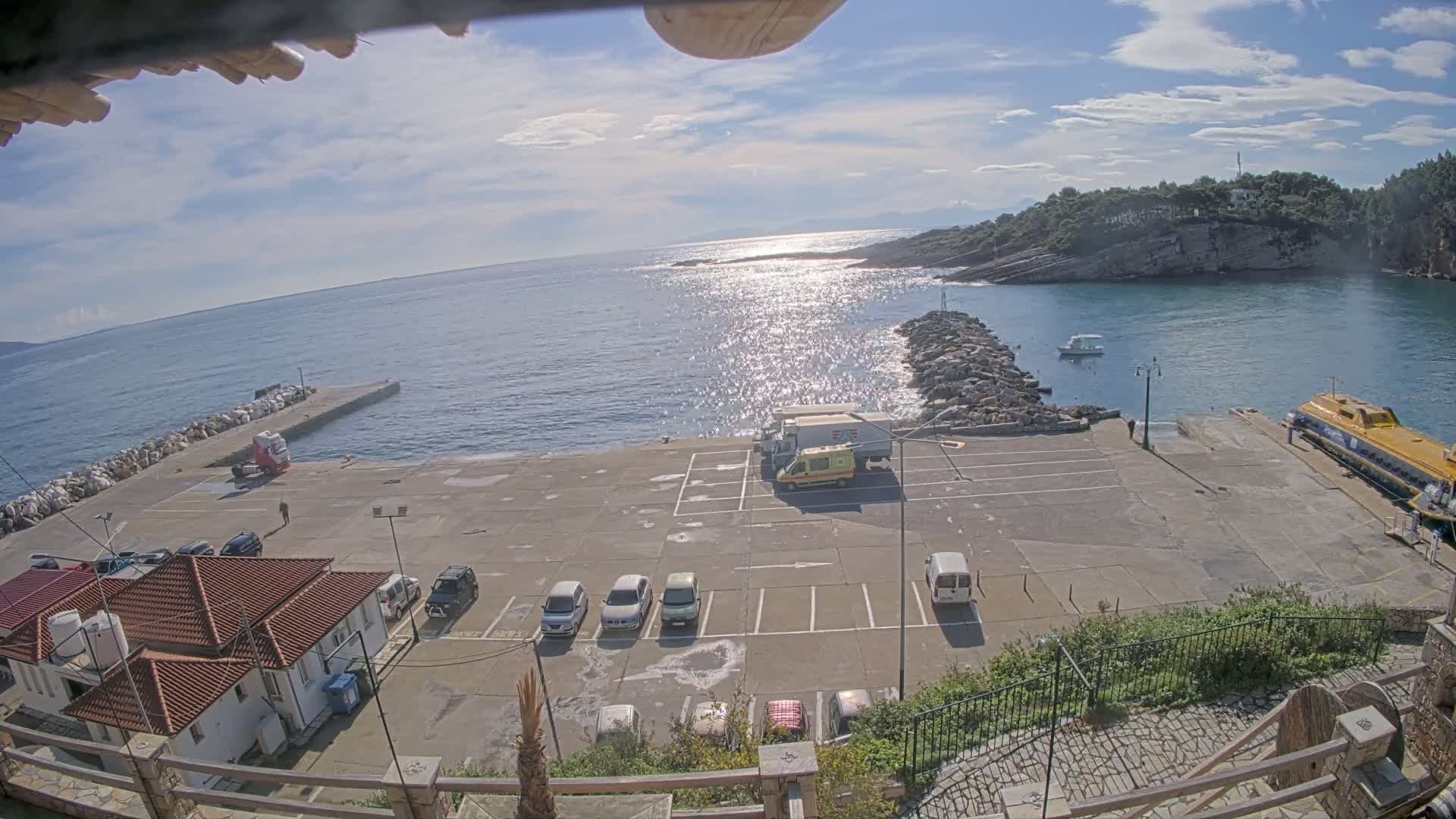 Under a partly cloudy sky with bright sunshine glinting on the blue sea, a harbor scene features a concrete pier filled with parked vehicles, a yellow catamaran docked, a small white boat on the water, and rocky, forested coastlines with buildings sporting terracotta roofs.