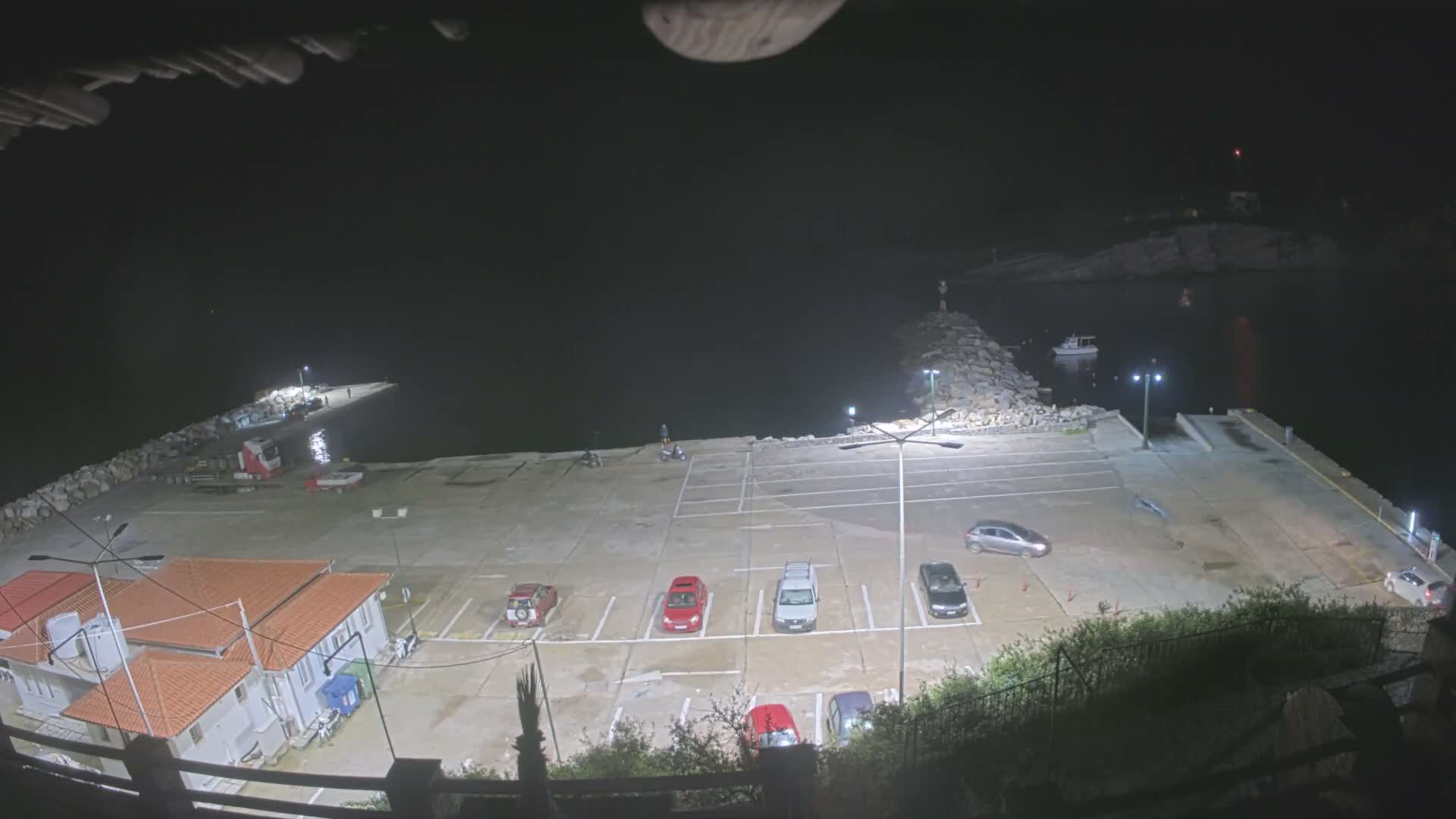 Under a partly cloudy sky with bright sunshine glinting on the blue sea, a harbor scene features a concrete pier filled with parked vehicles, a yellow catamaran docked, a small white boat on the water, and rocky, forested coastlines with buildings sporting terracotta roofs.