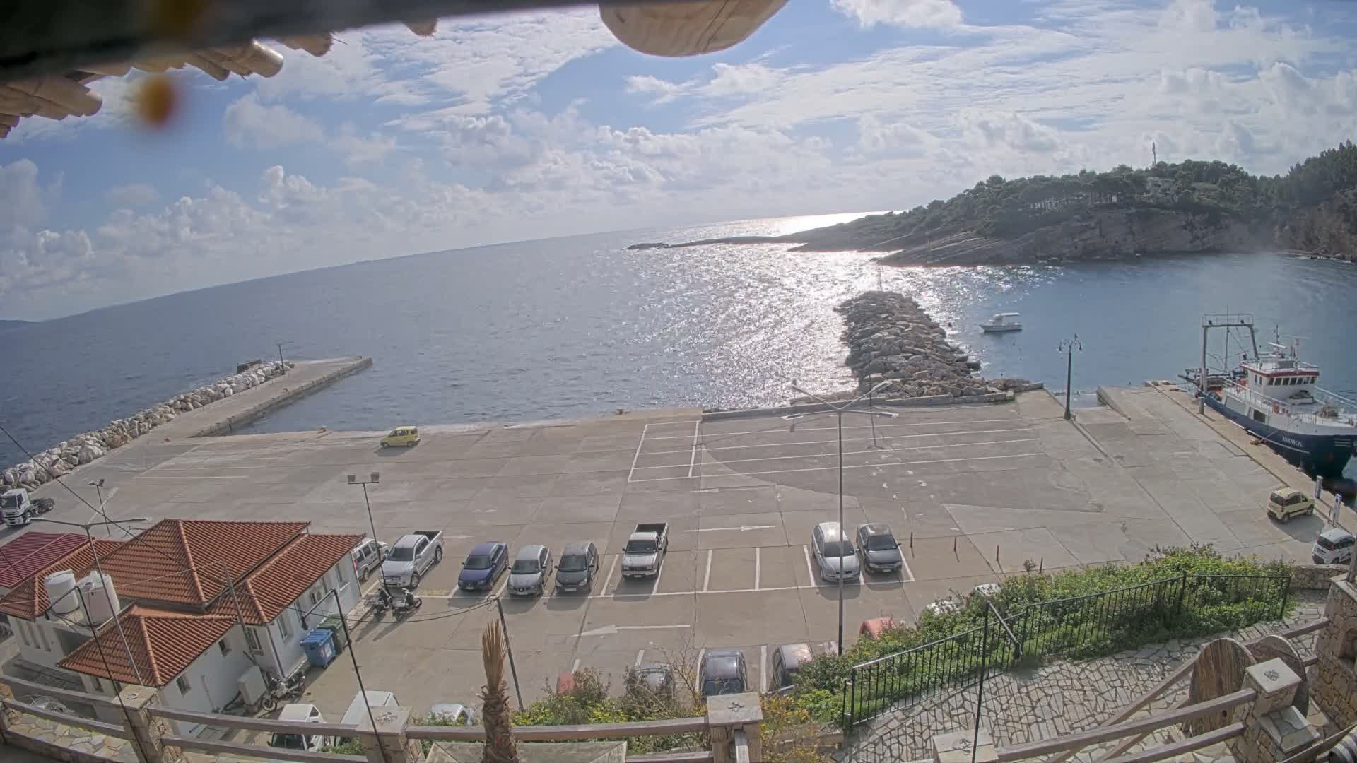 On a bright, partly cloudy day, a coastal harbor is visible with a large vessel docked at a pier, a parking lot containing multiple cars, buildings with orange tiled roofs, and the sea shimmering under the sun towards a distant, rocky island.