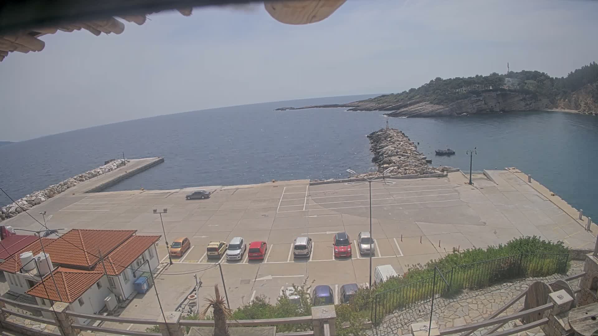 On a bright, partly cloudy day, a coastal harbor is visible with a large vessel docked at a pier, a parking lot containing multiple cars, buildings with orange tiled roofs, and the sea shimmering under the sun towards a distant, rocky island.