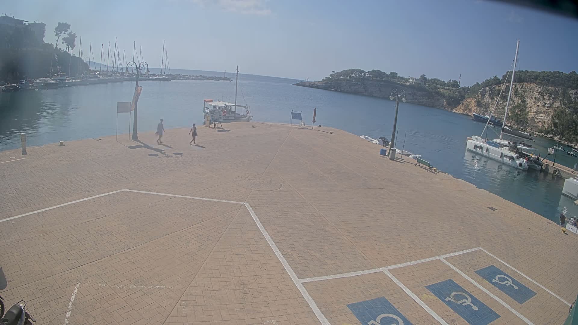 A paved plaza overlooks a calm harbor filled with boats on a sunny day.