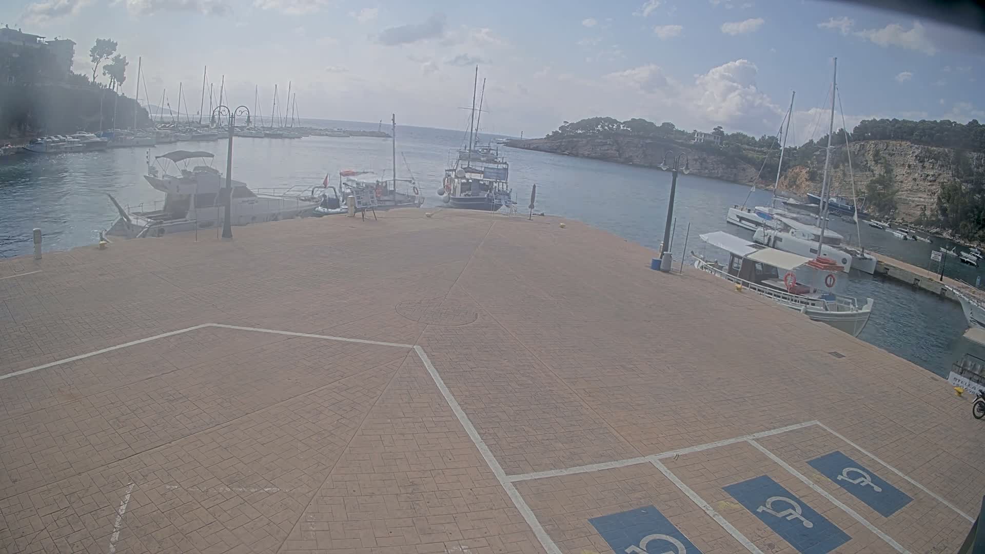 A paved area with several docked boats in a calm harbor under a partly cloudy sky.