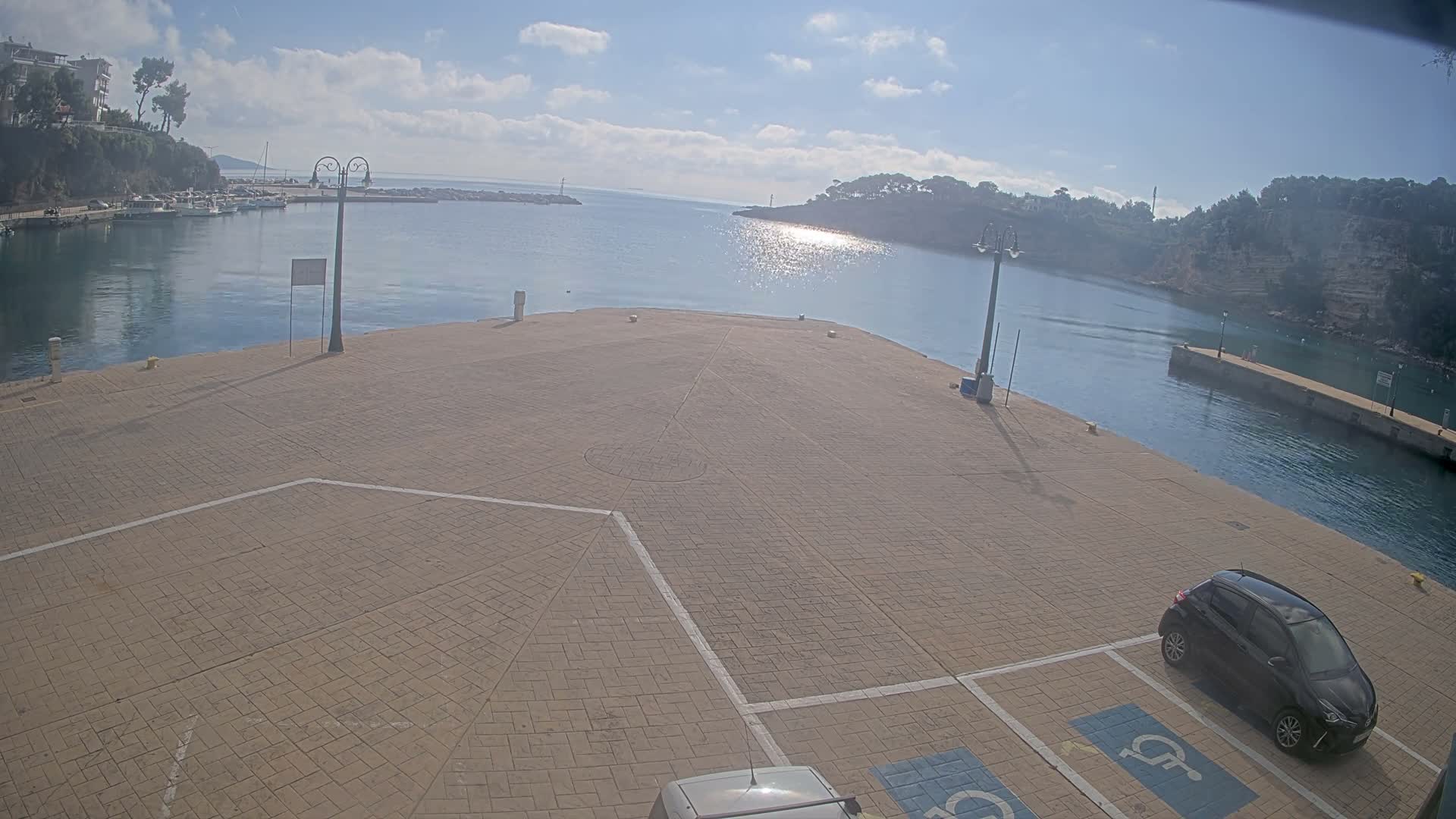 A bright, sunny day illuminates a paved harbor pier with a black car parked in the foreground, leading to calm blue water where sunlight glints, flanked by distant boats, tree-lined hills, and a sky with scattered clouds.