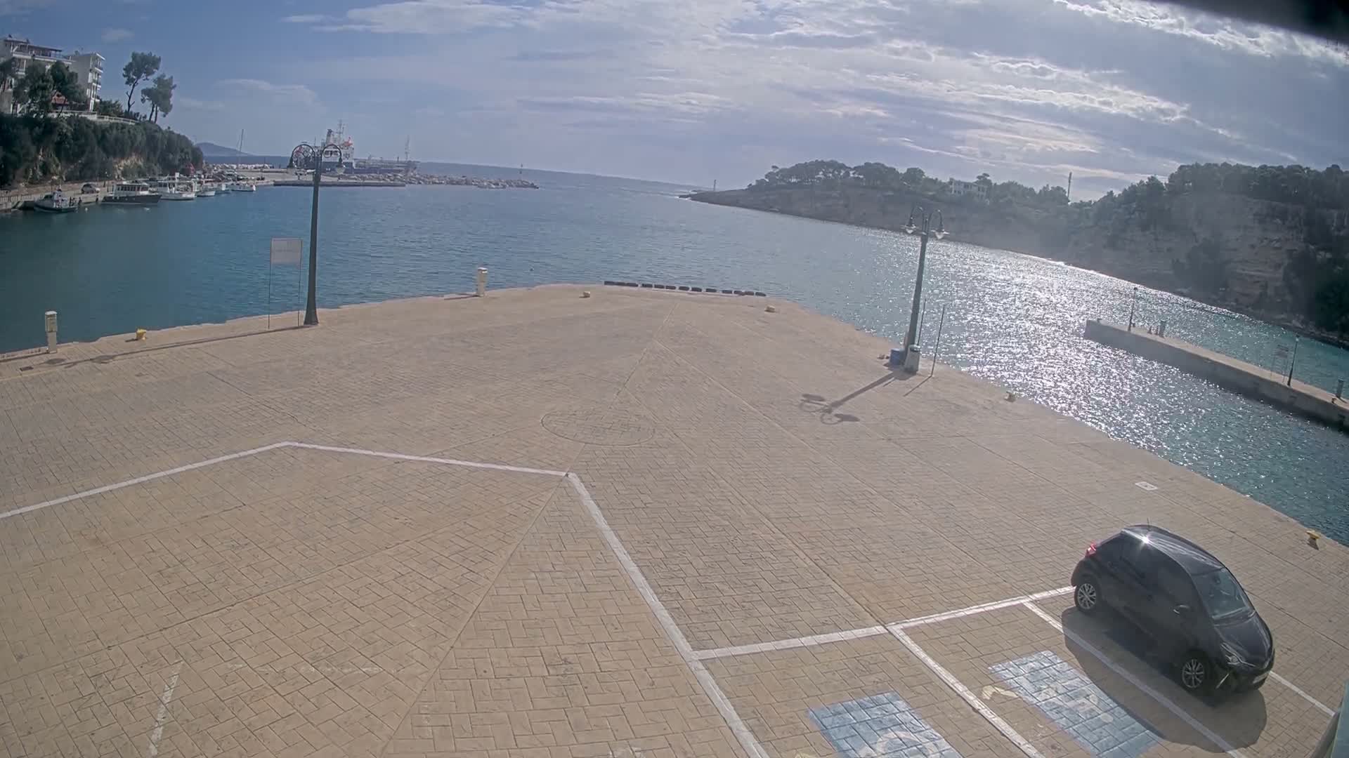 A bright, sunny day illuminates a paved harbor pier with a black car parked in the foreground, leading to calm blue water where sunlight glints, flanked by distant boats, tree-lined hills, and a sky with scattered clouds.