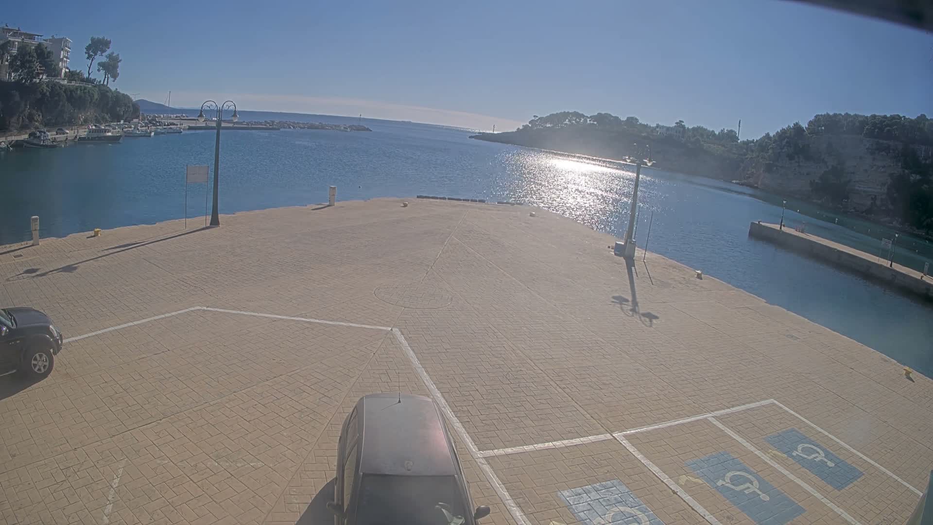 A bright, sunny day illuminates a wide paved pier and a bustling marina with boats docked on the left, leading to an open expanse of blue water reflecting the sun's glare, bordered by tree-covered hills under a clear sky.