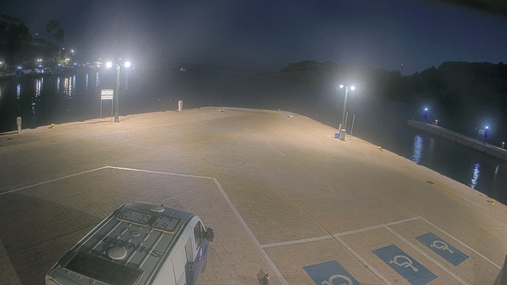 A well-lit pier or dock at night overlooks a calm body of water with distant illuminated shores, while a light-colored van is partially visible in the foreground on the paved surface marked with handicapped parking spots, all under a clear, dark sky.