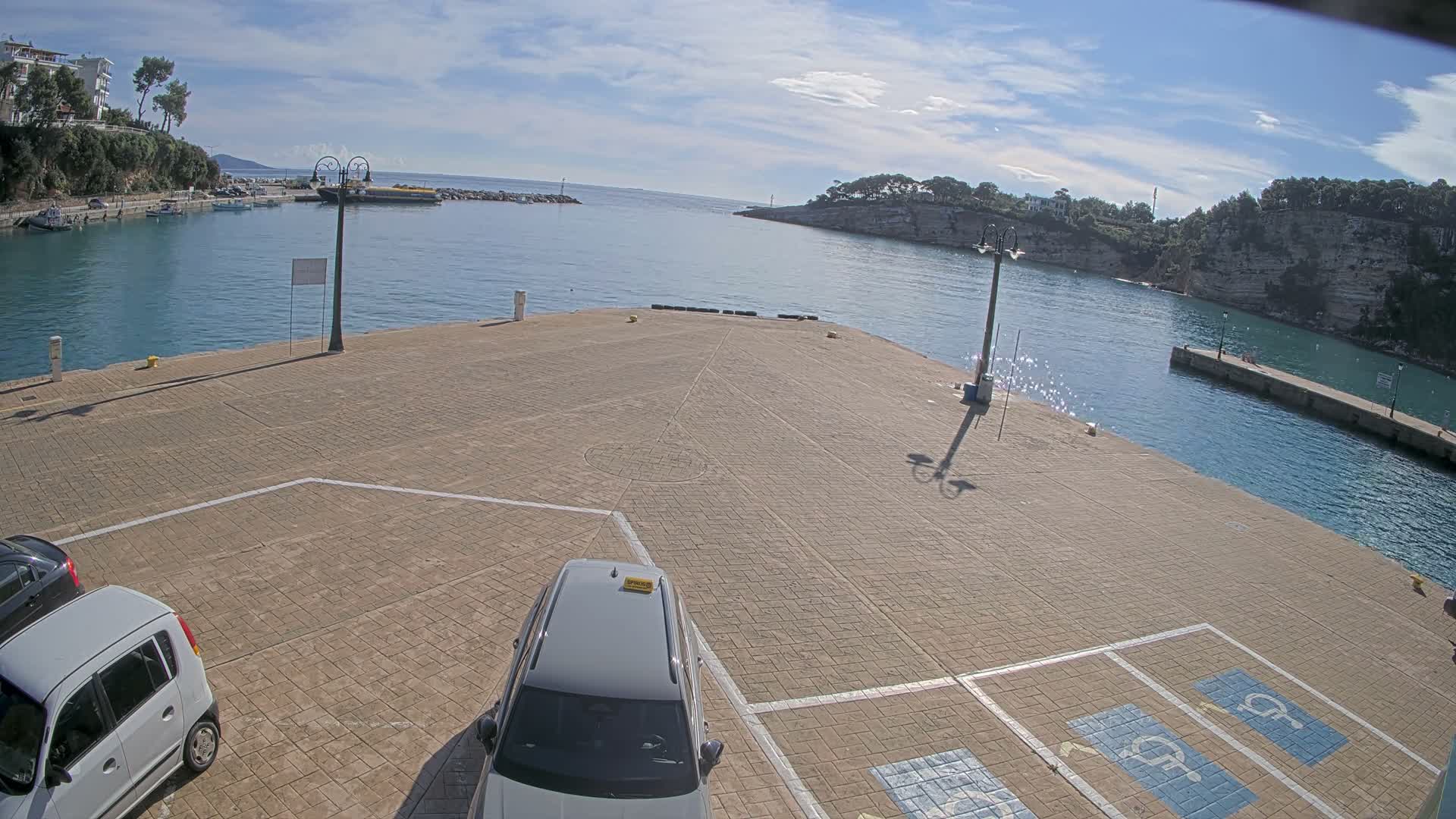 A sunny, wide-angle view of a tranquil harbor features a paved pier with two parked cars in the foreground, calm turquoise waters with boats docked along the left shore, and distant tree-lined coasts under a clear sky with scattered white clouds.