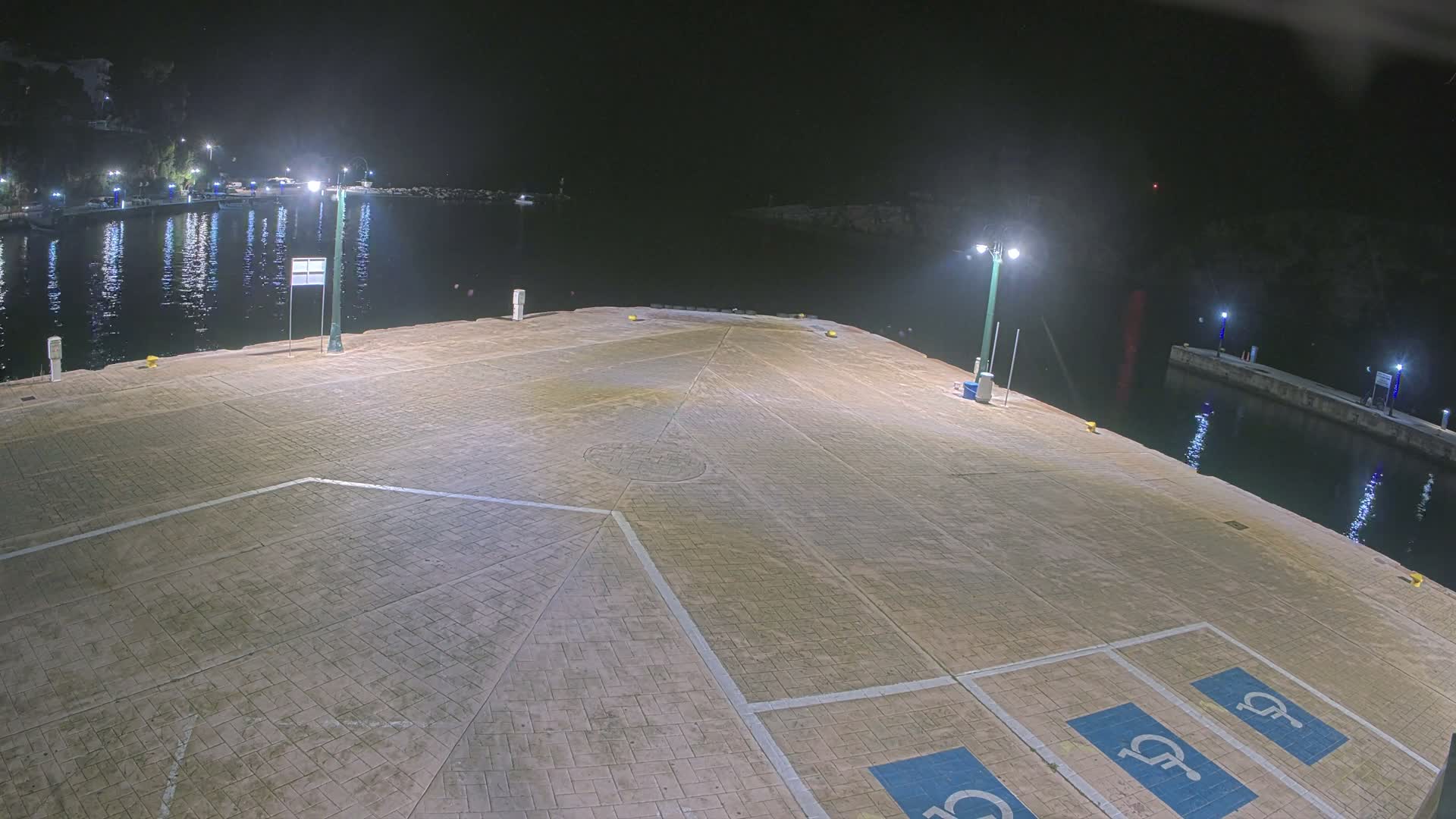 A sunny, wide-angle view of a tranquil harbor features a paved pier with two parked cars in the foreground, calm turquoise waters with boats docked along the left shore, and distant tree-lined coasts under a clear sky with scattered white clouds.
