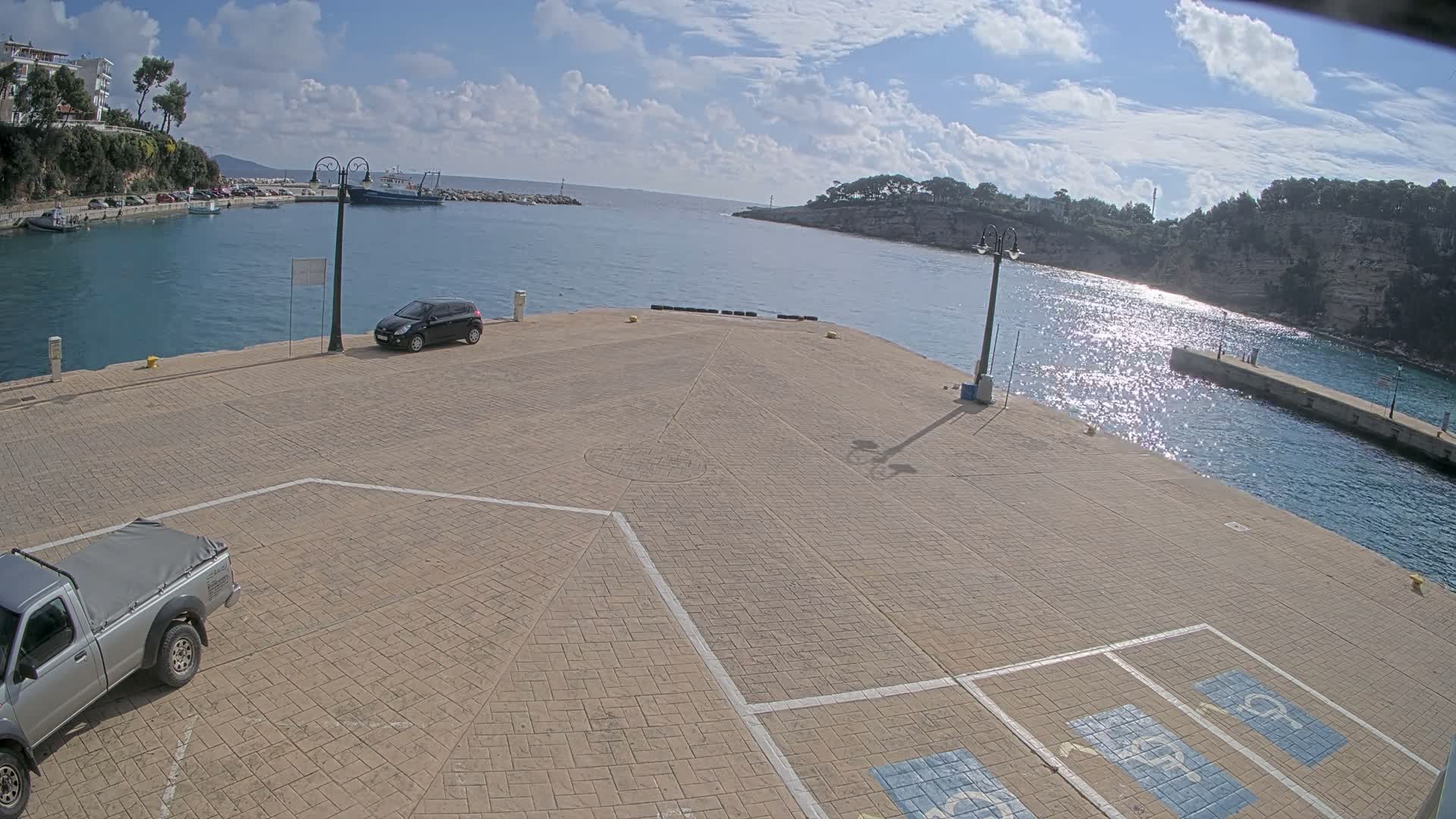 A bright, sunny day illuminates a tranquil harbor with sparkling blue waters and a wide paved pier featuring parked cars, all framed by distant green hills and a blue sky scattered with white clouds.