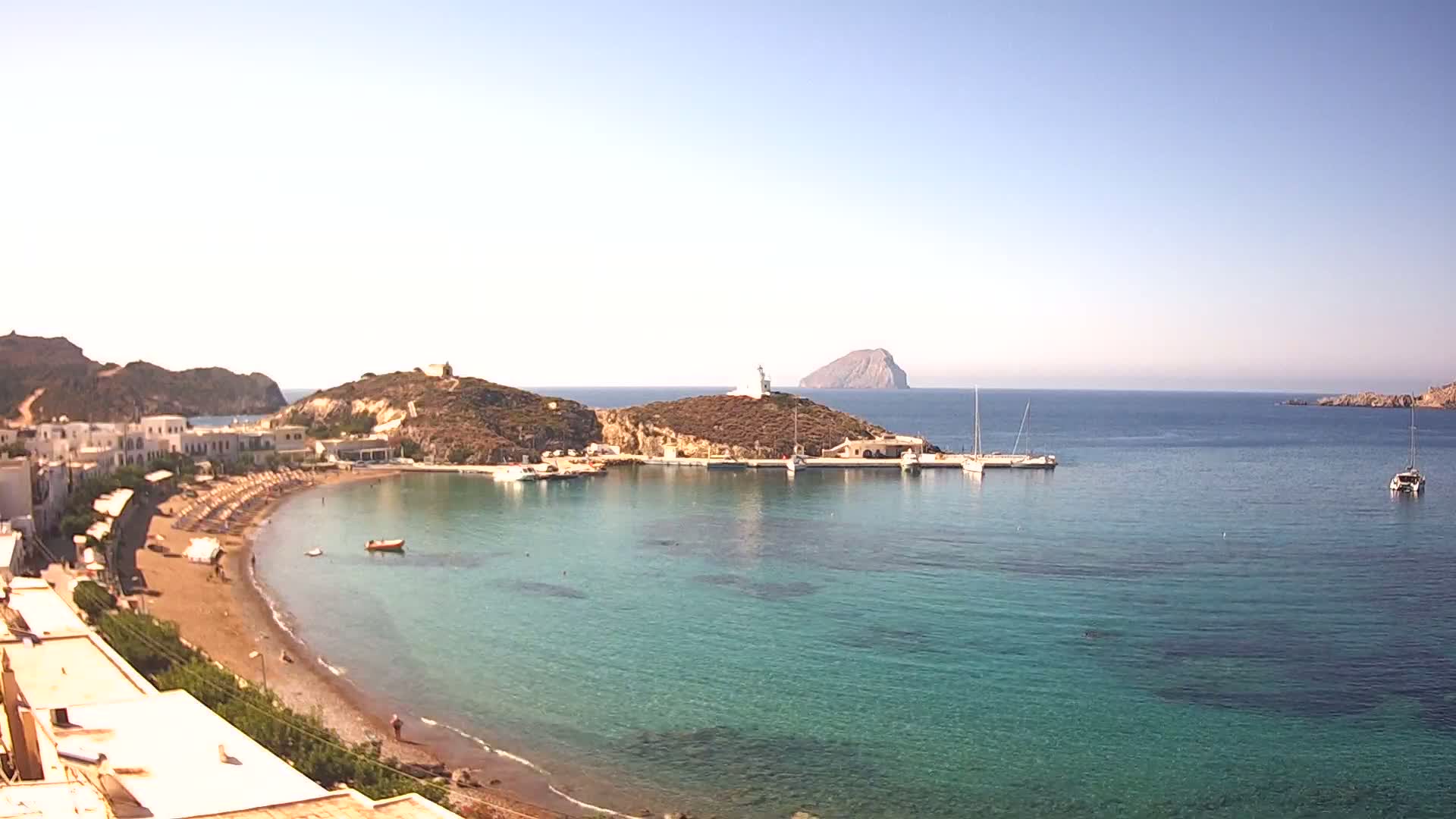 A sandy beach curves into a calm, clear turquoise bay with boats moored near a small town and a lighthouse on a nearby hill, under a bright sunny sky.