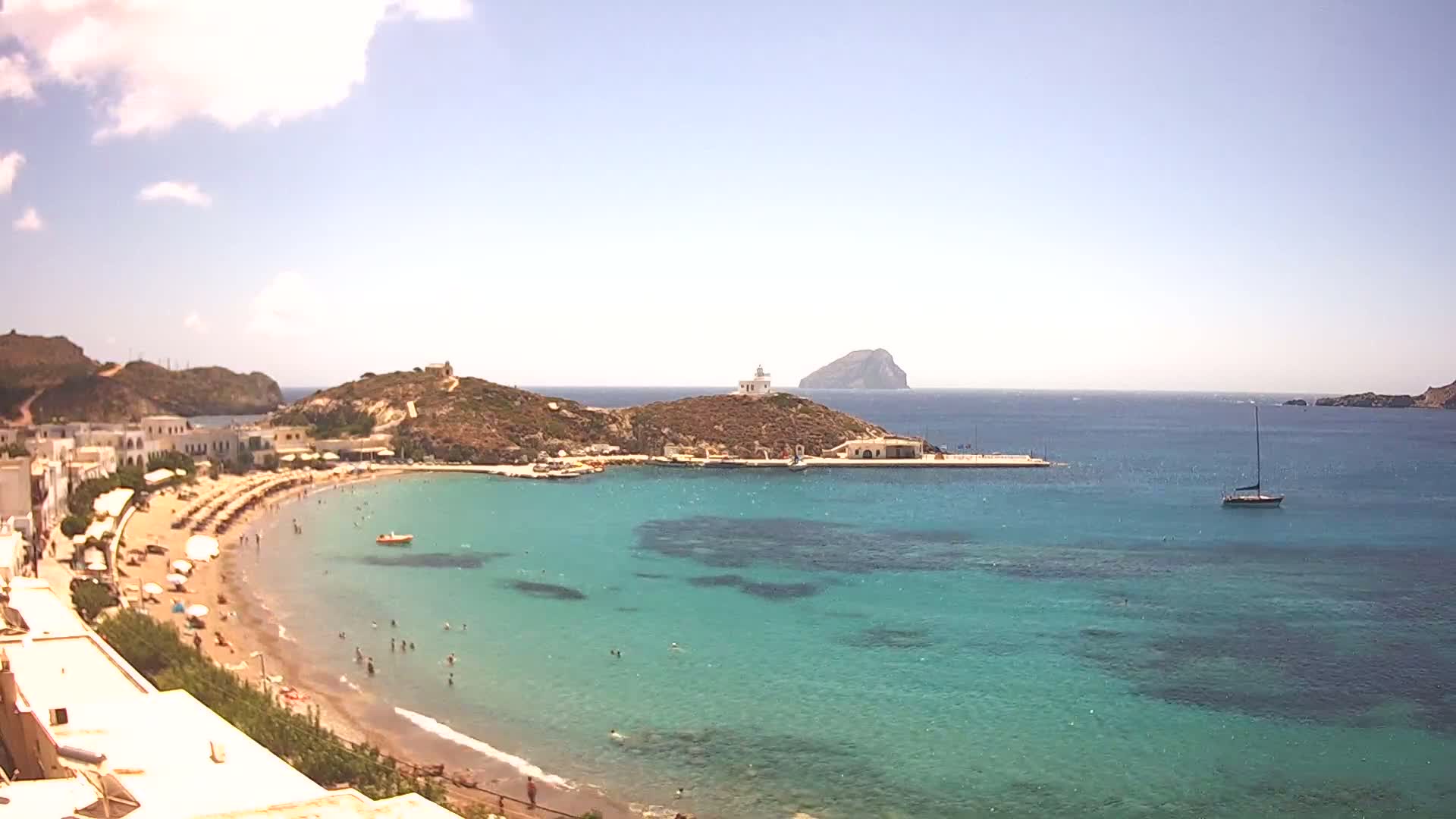 A sandy beach curves into a calm, clear turquoise bay with boats moored near a small town and a lighthouse on a nearby hill, under a bright sunny sky.