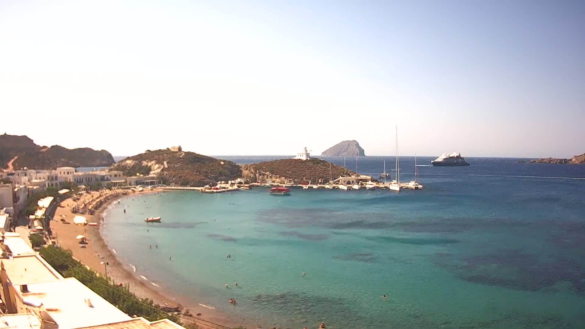 A sunny day shows a small town's curved sandy beach and calm turquoise waters, with boats in a harbor and a large ship in the distance, all nestled between hills and islands.