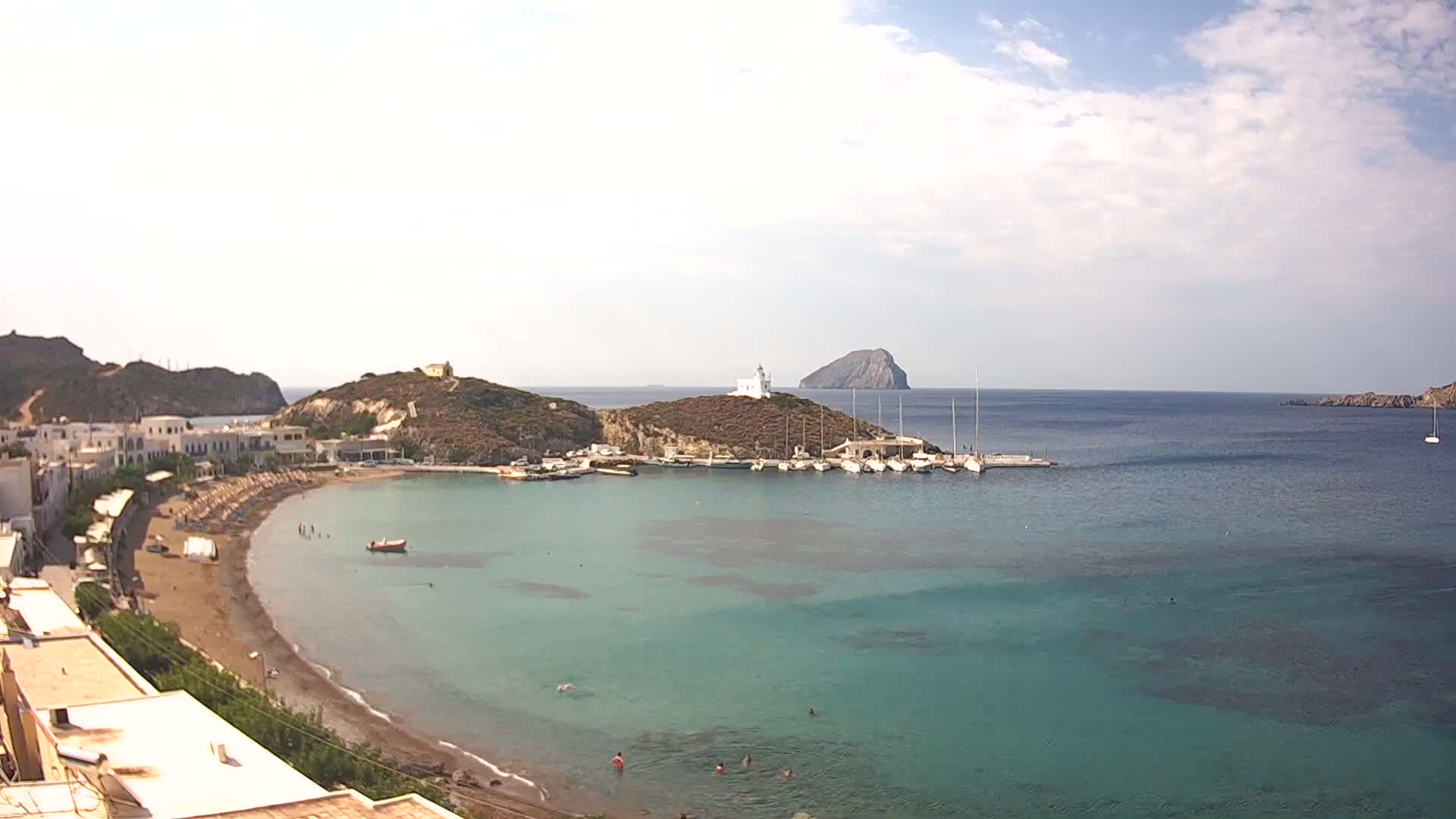 A small, sandy beach curves into a calm, turquoise bay, where several boats are moored near a white lighthouse situated on a small, hilly island under a partly cloudy sky.