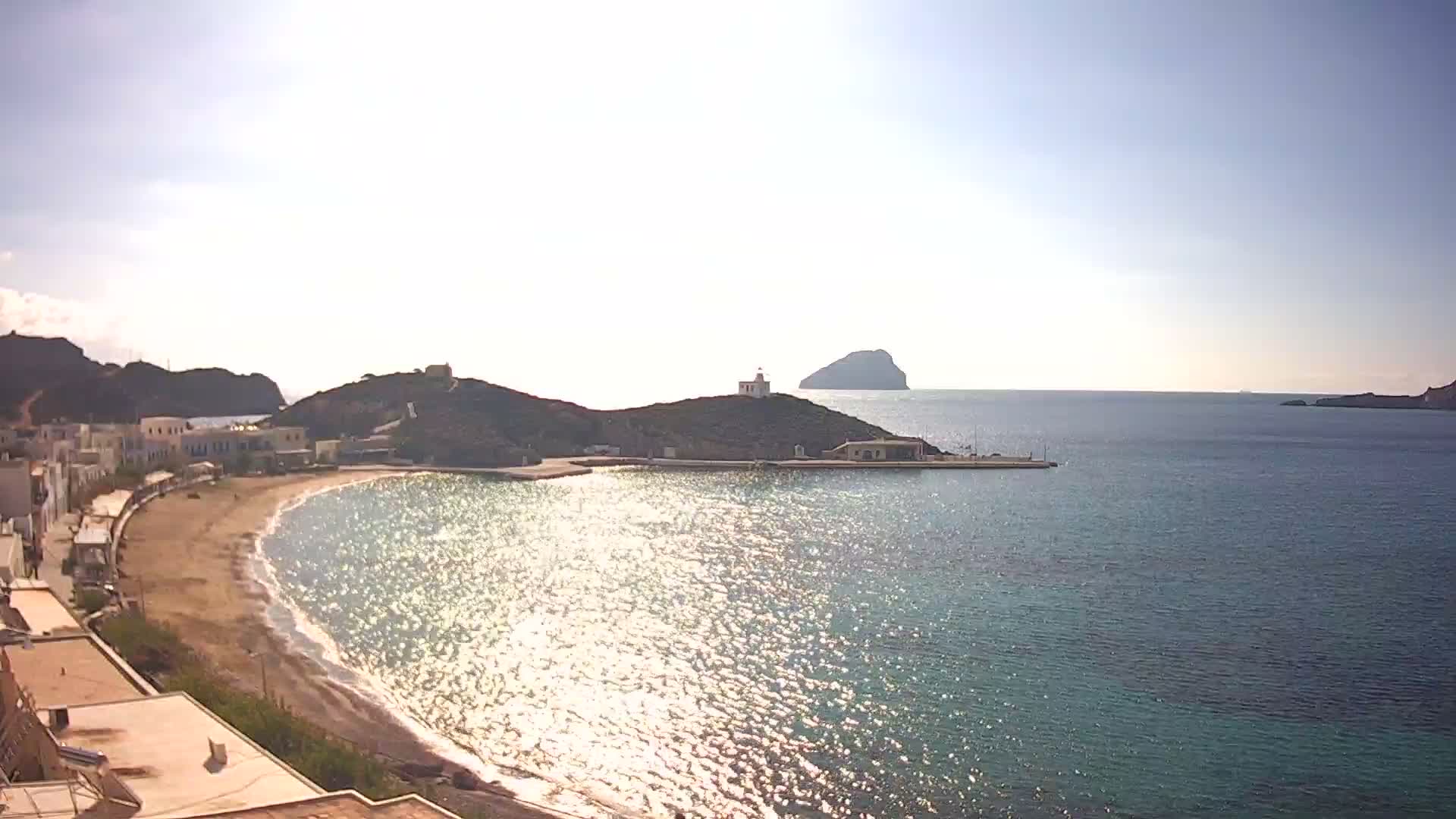 A bright and sunny day reveals a picturesque coastal village with a sandy beach curving along a bay, overlooked by a lighthouse on a hill and a small island in the sparkling blue sea.