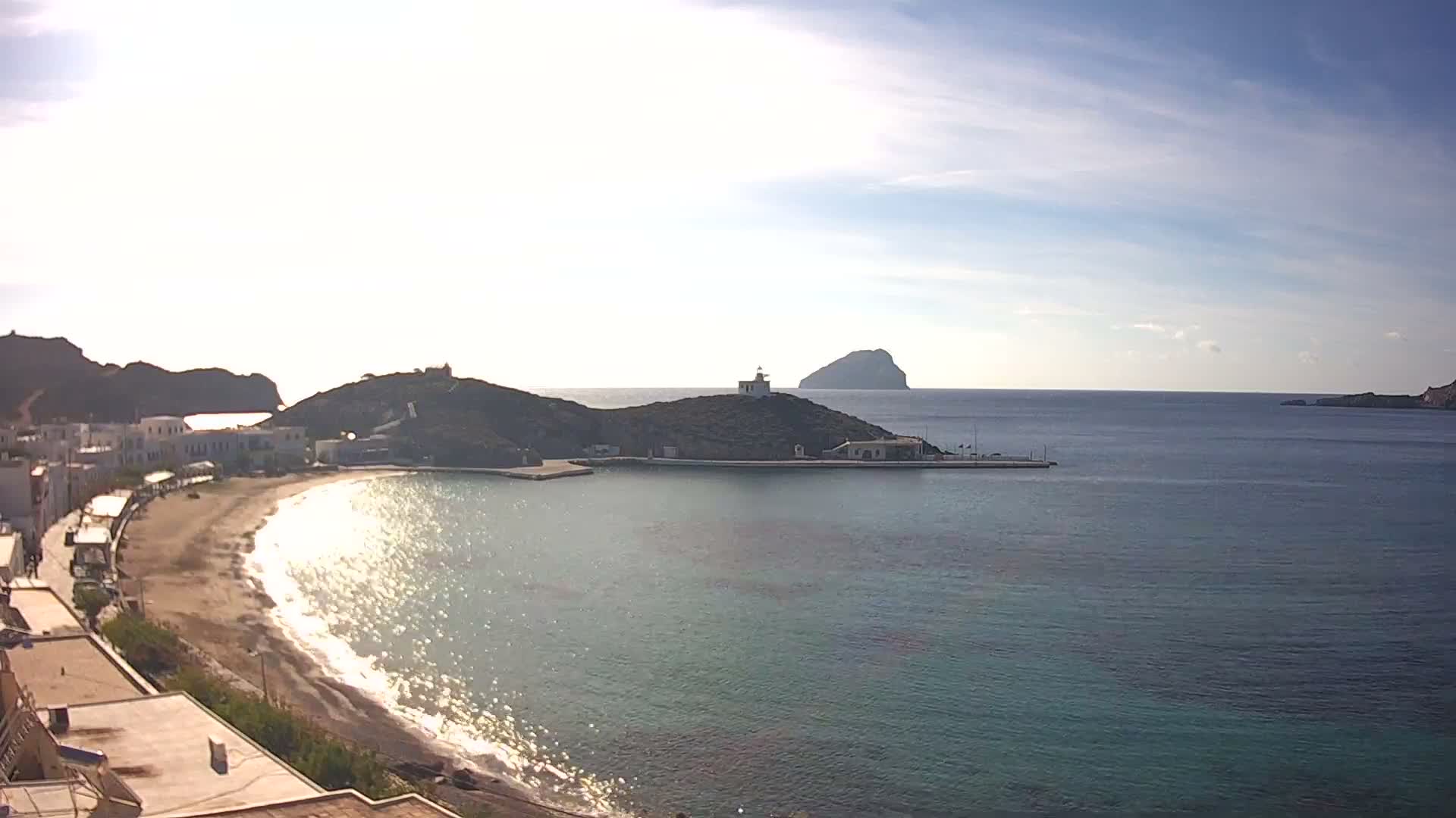 A bright, sunny day illuminates a coastal town nestled by a curving sandy beach, with a bay featuring a lighthouse on a prominent hill, a small pier, and a distant island under a partly cloudy sky.