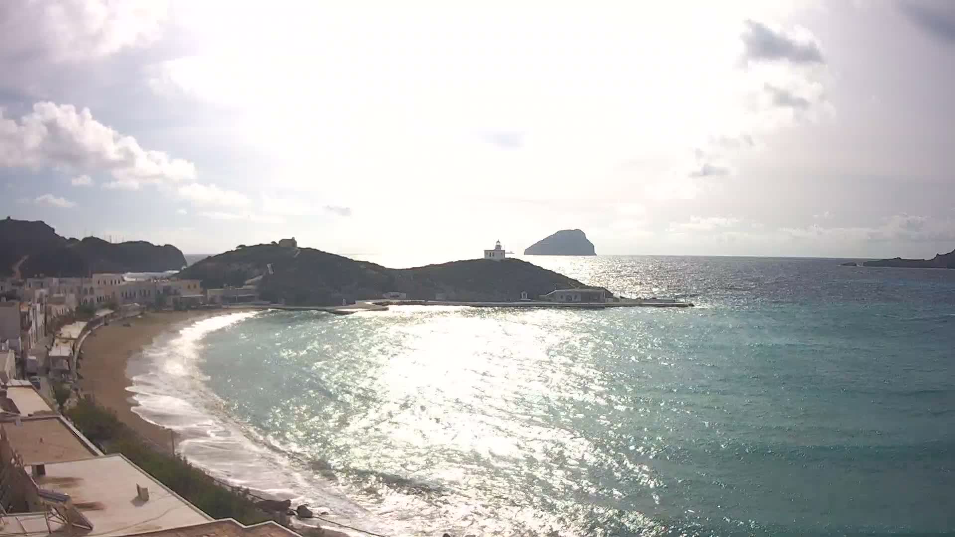 A bright, sunny day illuminates a coastal town nestled by a curving sandy beach, with a bay featuring a lighthouse on a prominent hill, a small pier, and a distant island under a partly cloudy sky.