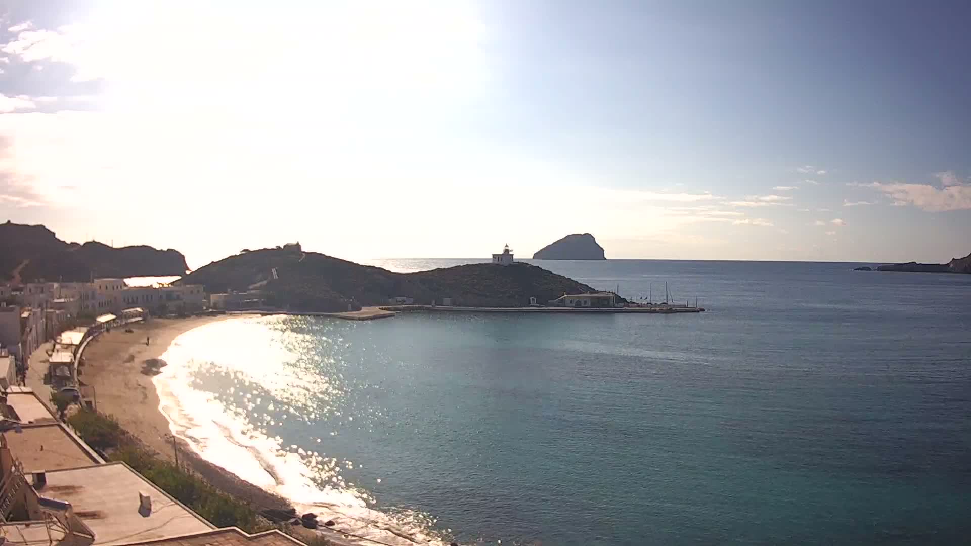 A bright, sunny day illuminates a coastal village with white buildings alongside a sandy beach, a sparkling blue bay, and a lighthouse perched on a green peninsula, with a distant island on the horizon under a clear sky.