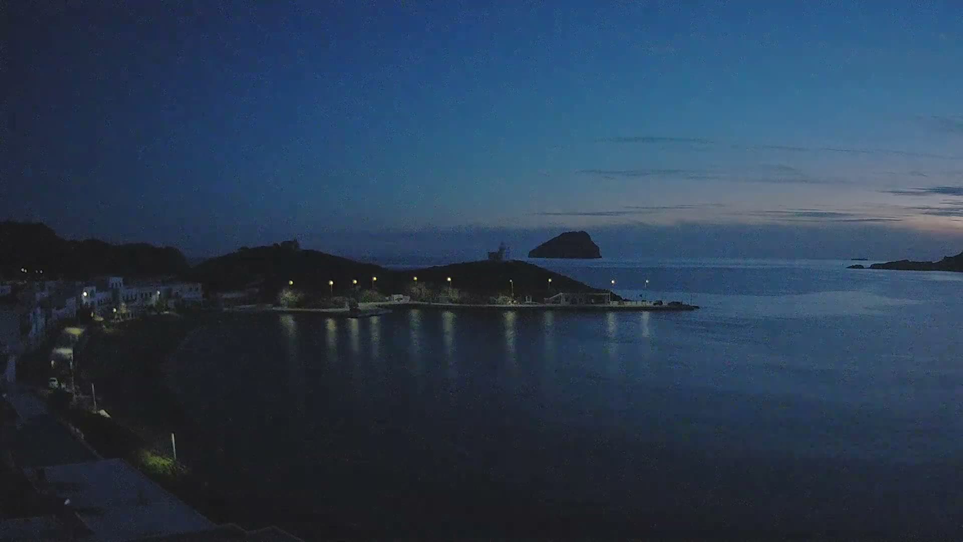 A tranquil coastal bay is illuminated by the streetlights and buildings of a town along its shore, reflecting on the calm water under a clear to partly cloudy dark blue sky, with dark hills and a small island visible in the distance.