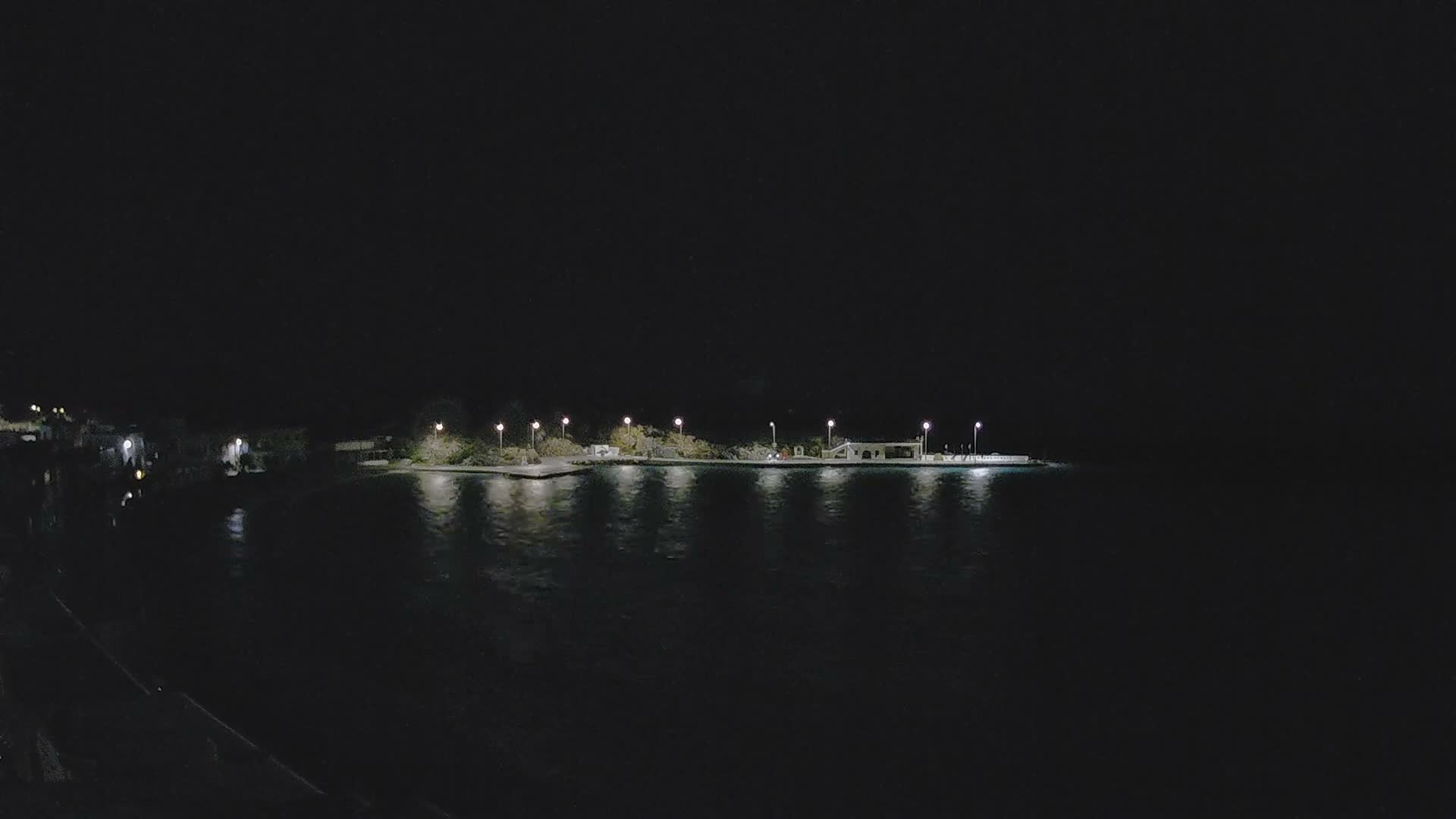 A tranquil coastal bay is illuminated by the streetlights and buildings of a town along its shore, reflecting on the calm water under a clear to partly cloudy dark blue sky, with dark hills and a small island visible in the distance.