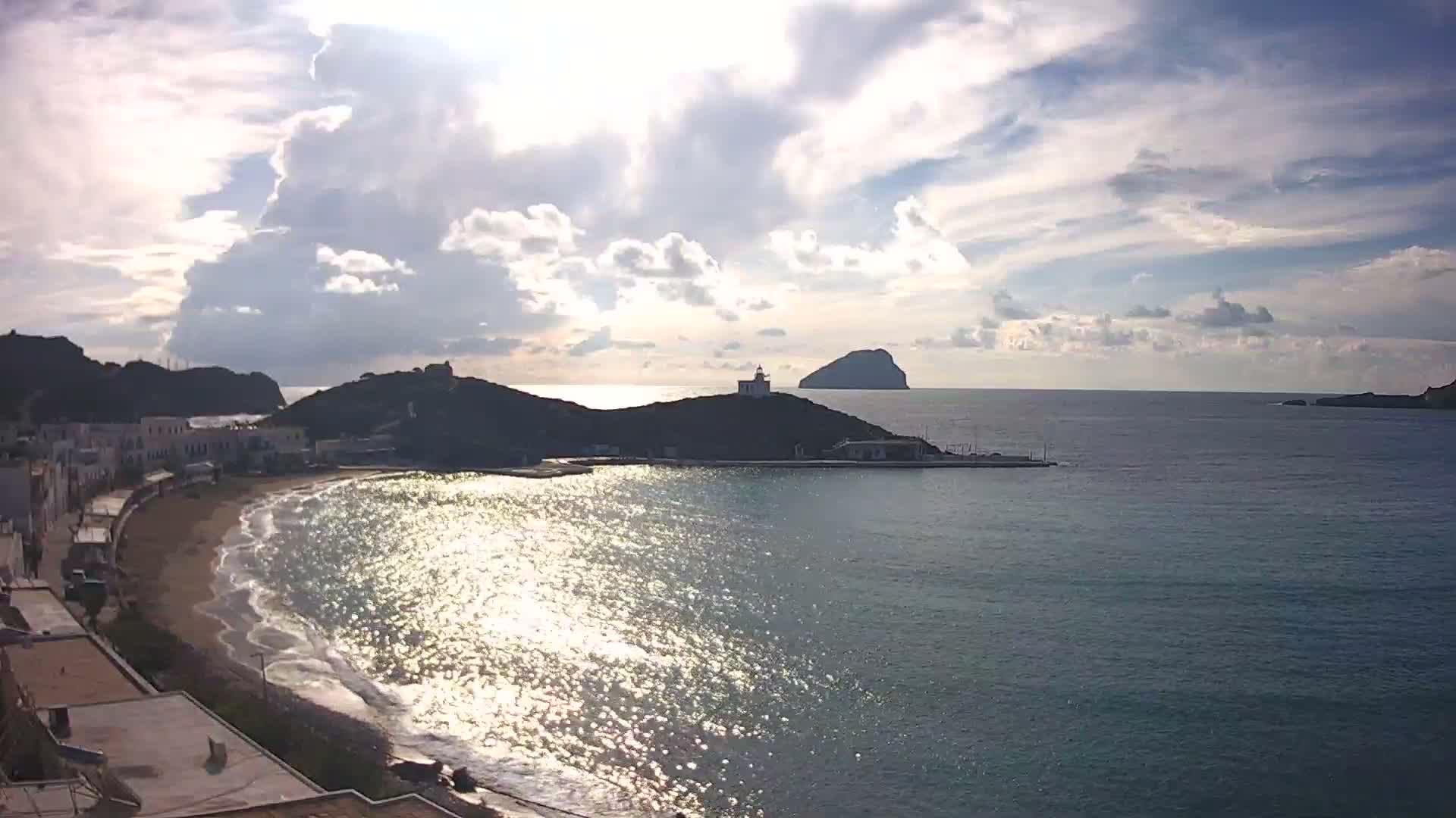 A bright, partly cloudy sky hangs over a coastal town featuring a crescent beach and buildings, facing a sparkling sea with a lighthouse on a nearby hill and a distant island.