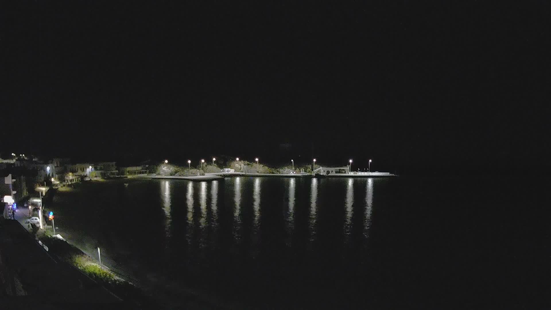 A bright, partly cloudy sky hangs over a coastal town featuring a crescent beach and buildings, facing a sparkling sea with a lighthouse on a nearby hill and a distant island.