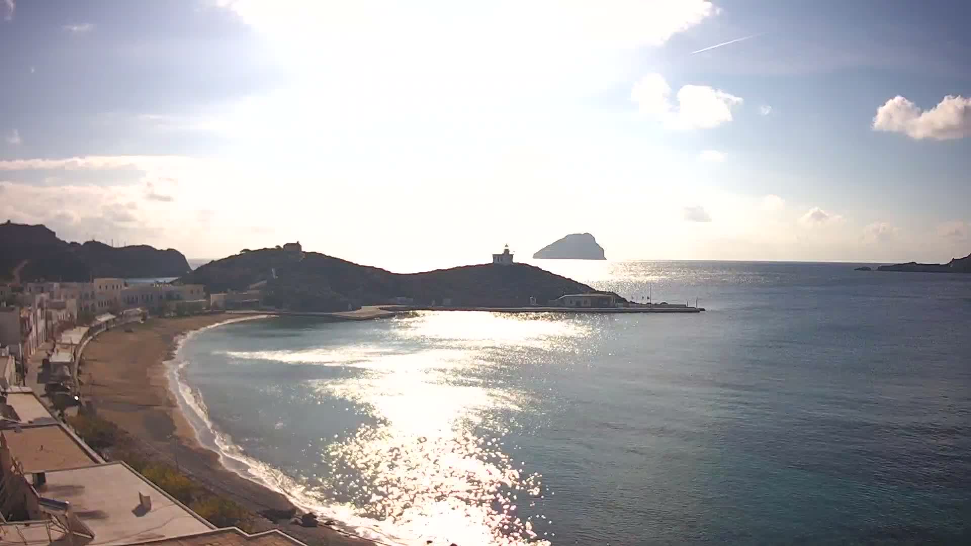 A bright and sunny coastal scene displays a town with white buildings along a crescent beach and calm, sparkling blue water, featuring a lighthouse atop a prominent hill and a distant island under a partly cloudy sky.