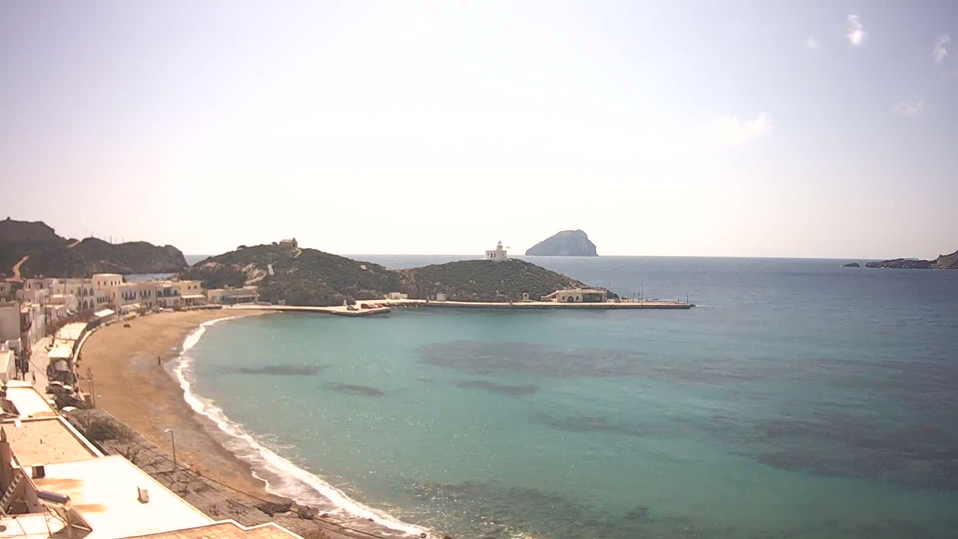 A bright and sunny coastal scene displays a town with white buildings along a crescent beach and calm, sparkling blue water, featuring a lighthouse atop a prominent hill and a distant island under a partly cloudy sky.