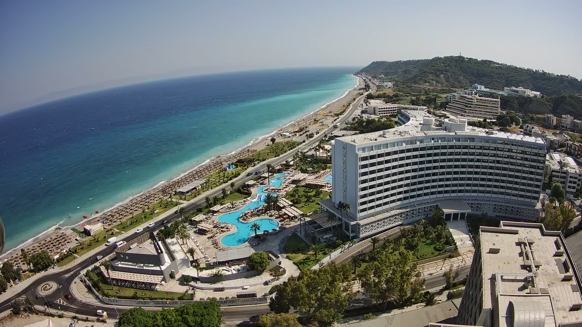 An aerial view shows a large coastal hotel complex with multiple pools and a beach lined with umbrellas under a clear, sunny sky.