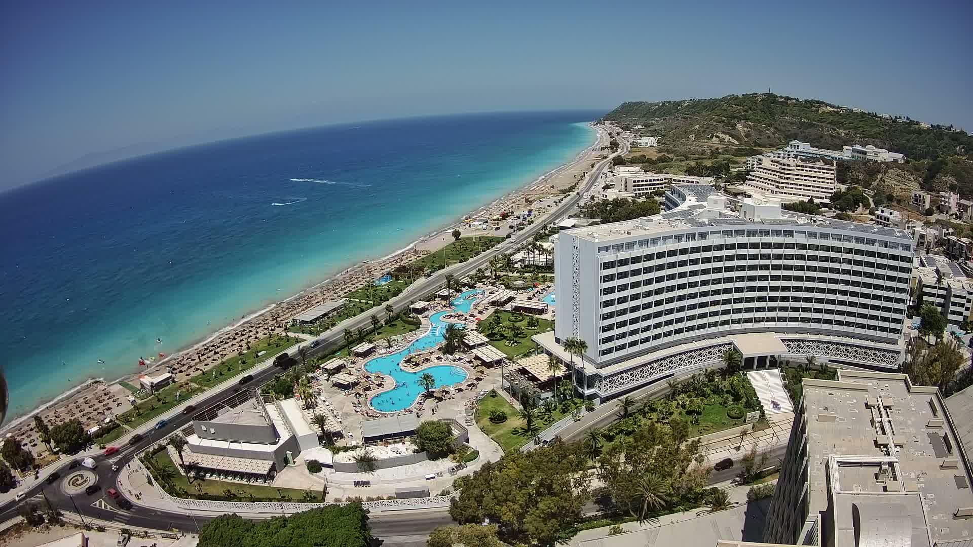 An aerial view shows a large coastal hotel complex with multiple pools and a beach lined with umbrellas under a clear, sunny sky.