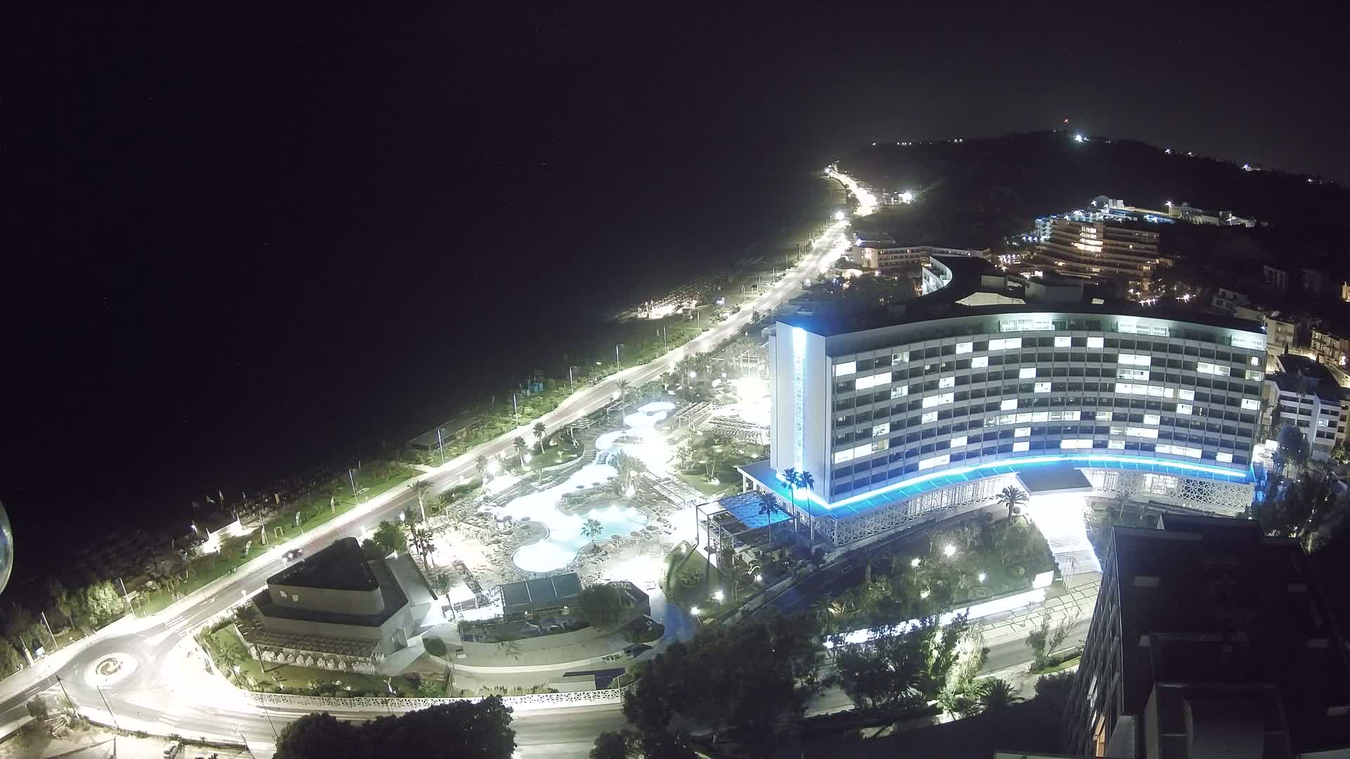 An aerial view shows a large coastal hotel complex with multiple pools and a beach lined with umbrellas under a clear, sunny sky.