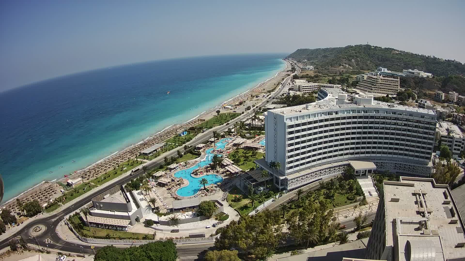 An aerial view shows a large hotel complex with multiple pools situated next to a sandy beach and turquoise ocean on a clear, sunny day.
