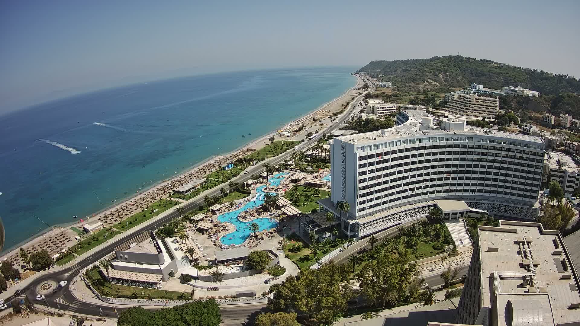 An aerial view shows a large resort hotel complex with multiple pools next to a sandy beach and clear blue ocean on a sunny day.