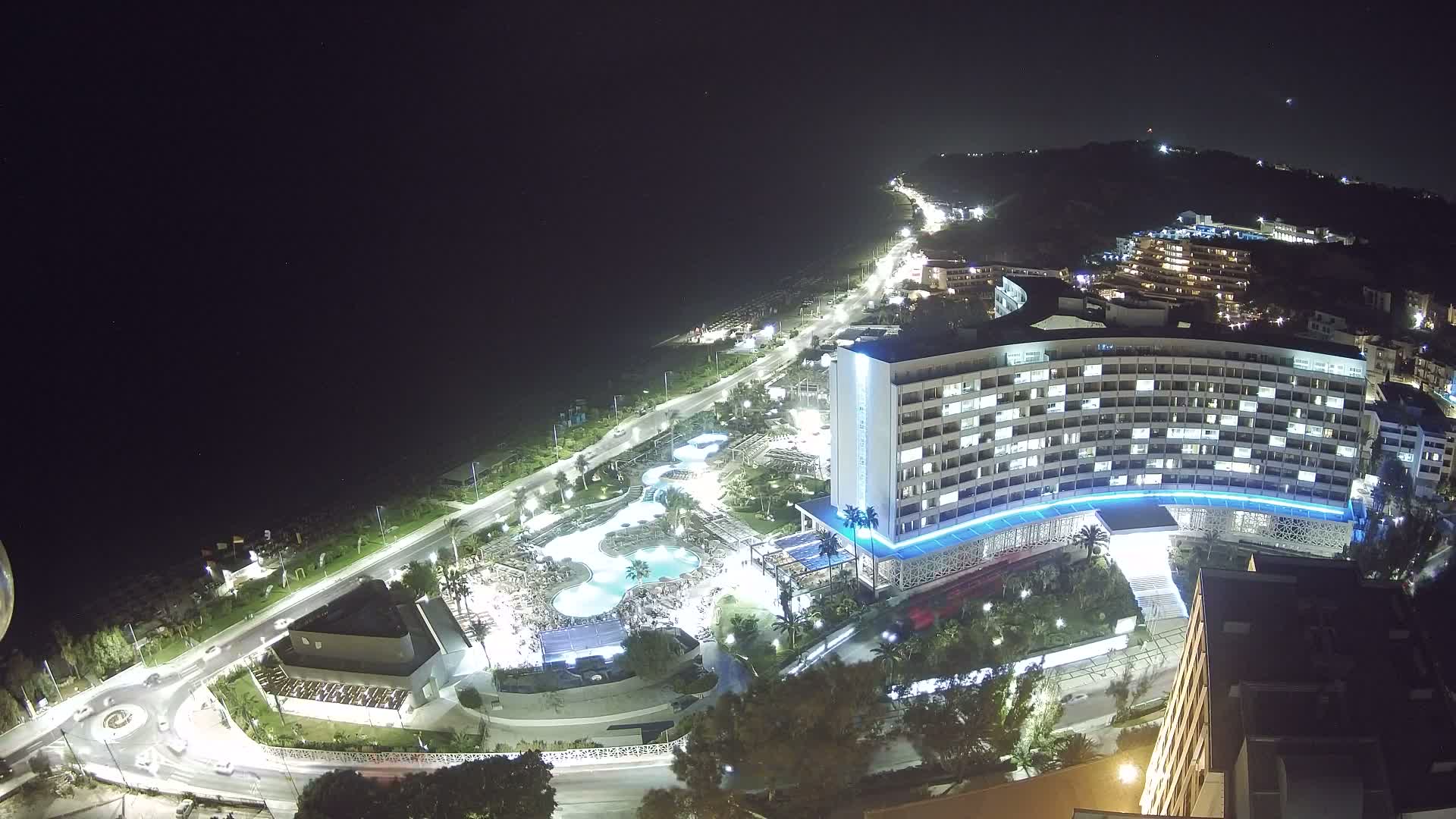 A large, brightly lit hotel complex sits near a coastline road at night, with surrounding buildings and pools also illuminated.