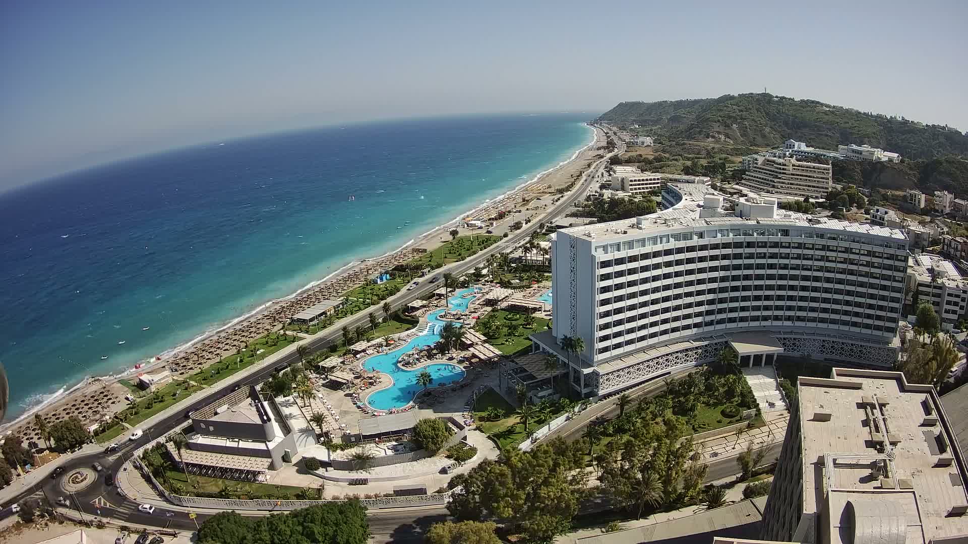 An aerial view shows a large beachfront hotel with multiple pools, surrounded by lush landscaping and a sandy beach with numerous umbrellas under a clear, sunny sky.