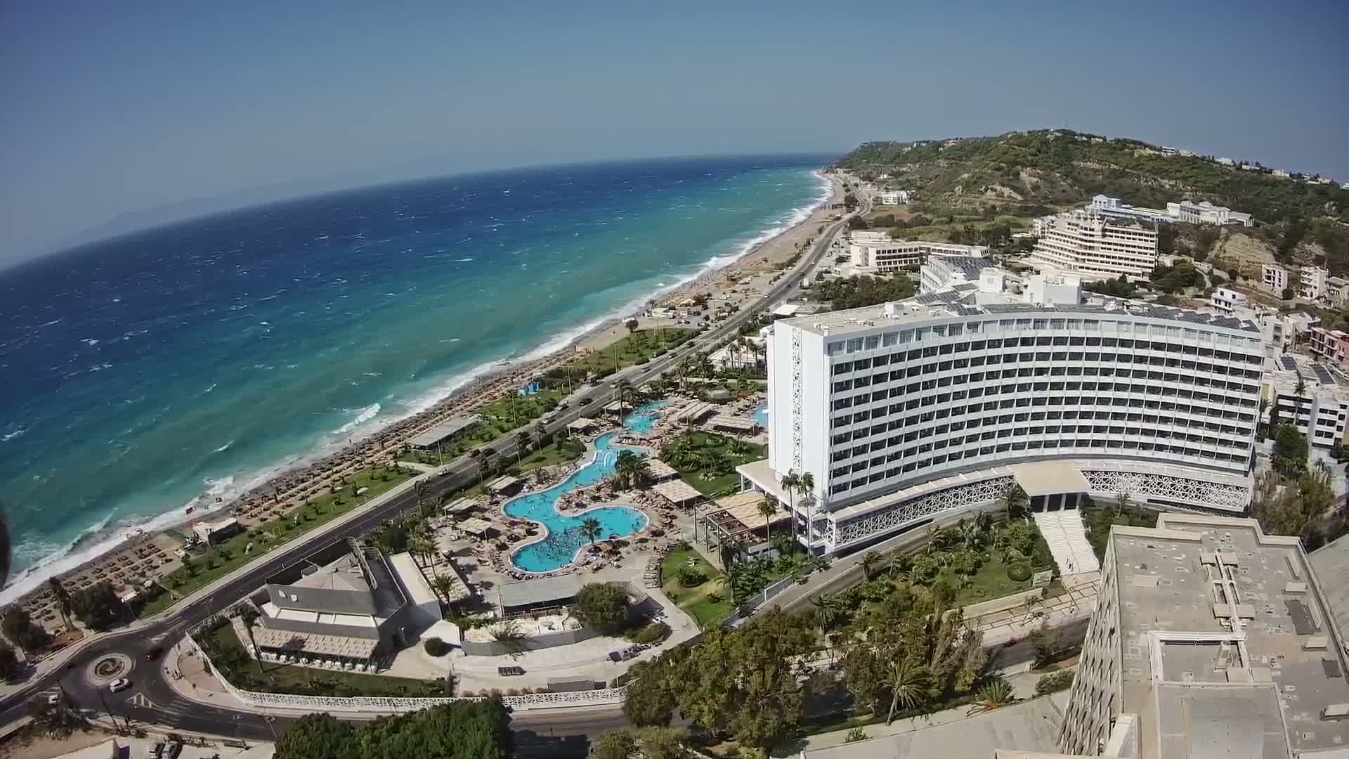 An aerial view shows a large beachfront hotel with multiple pools, surrounded by lush landscaping and a sandy beach with numerous umbrellas under a clear, sunny sky.