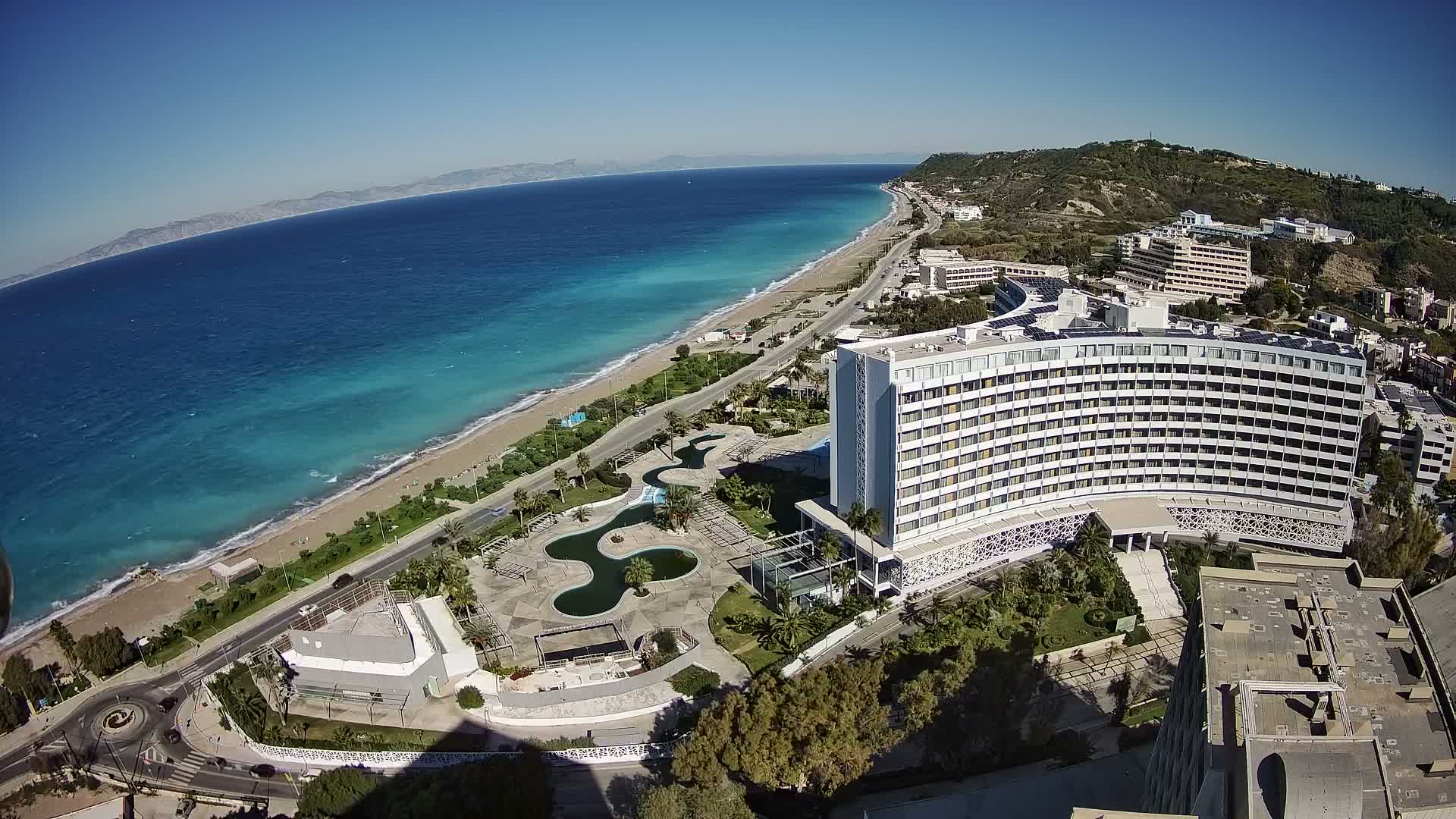 An aerial view reveals a sunny coastal landscape featuring a long sandy beach bordered by a multi-story white hotel, a curving road, and lush hills, with the clear blue ocean stretching towards distant mountains under a bright blue sky.